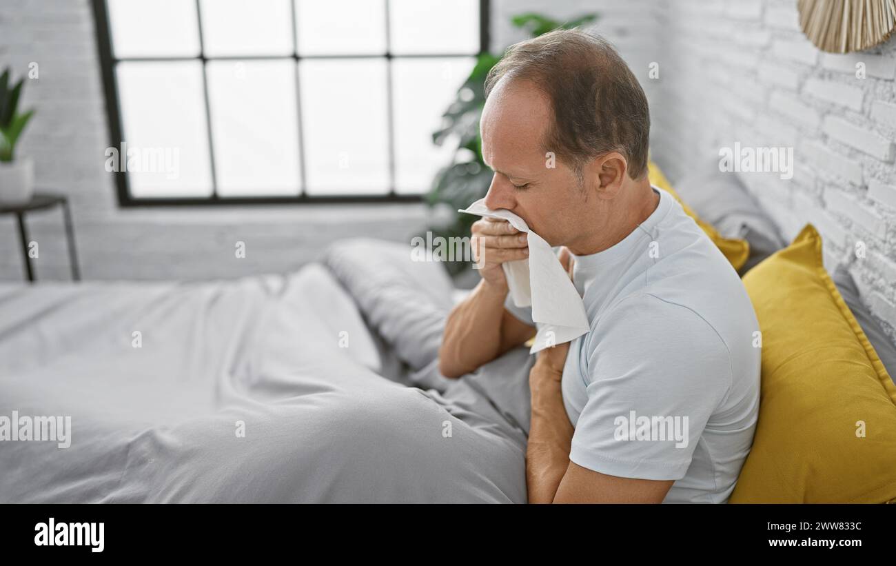 Un uomo di mezza età stressato soffre di malattia, seduto a letto in lutto nella camera da letto del suo appartamento Foto Stock