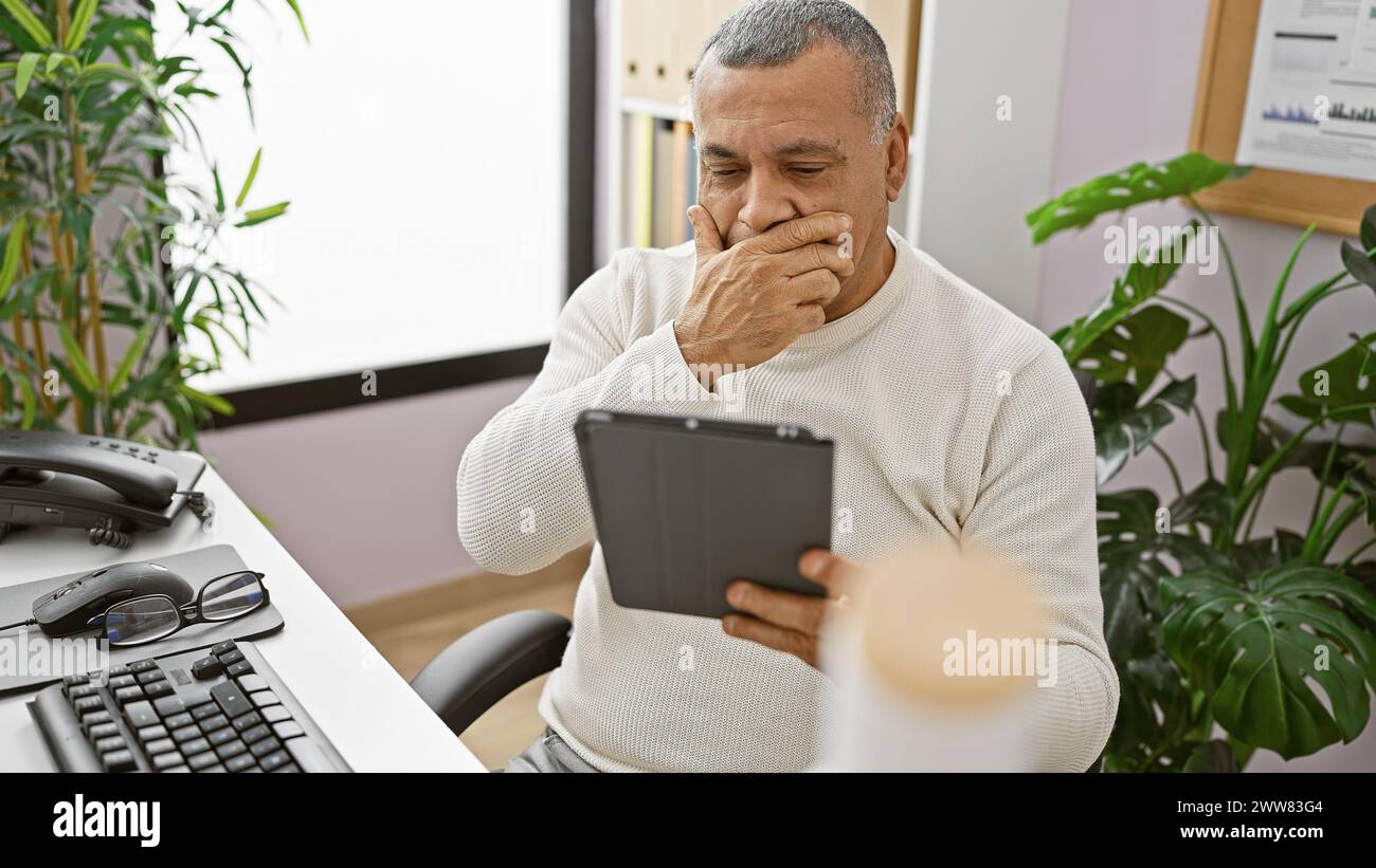 Un uomo ispanico maturo con un maglione bianco usa un tablet in un ufficio moderno, mostrando preoccupazione o pensiero profondo. Foto Stock