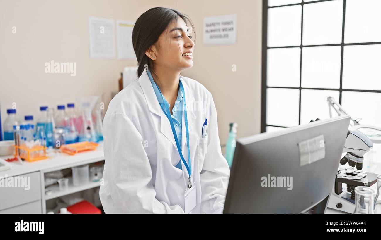 Una giovane scienziata indiana sorridente mentre lavora in un laboratorio al chiuso con un microscopio e un computer. Foto Stock