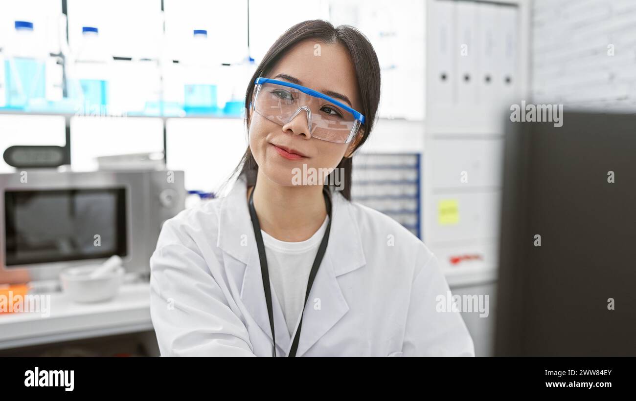 Una giovane donna asiatica che indossa un camice da laboratorio e occhiali di sicurezza si trova in un ambiente di laboratorio. Foto Stock