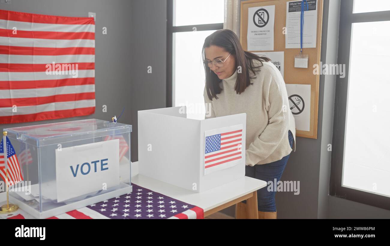 Una donna ispanica di mezza età vota al chiuso in un centro elettorale degli stati uniti con una bandiera americana visibile. Foto Stock