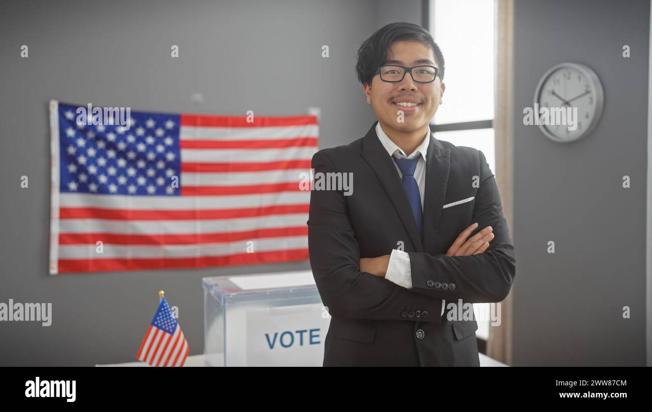 Sorridente asiatico con le braccia incrociate in piedi in un interno moderno con una bandiera US e una cabina di voto, che irradia fiducia e dovere civico. Foto Stock