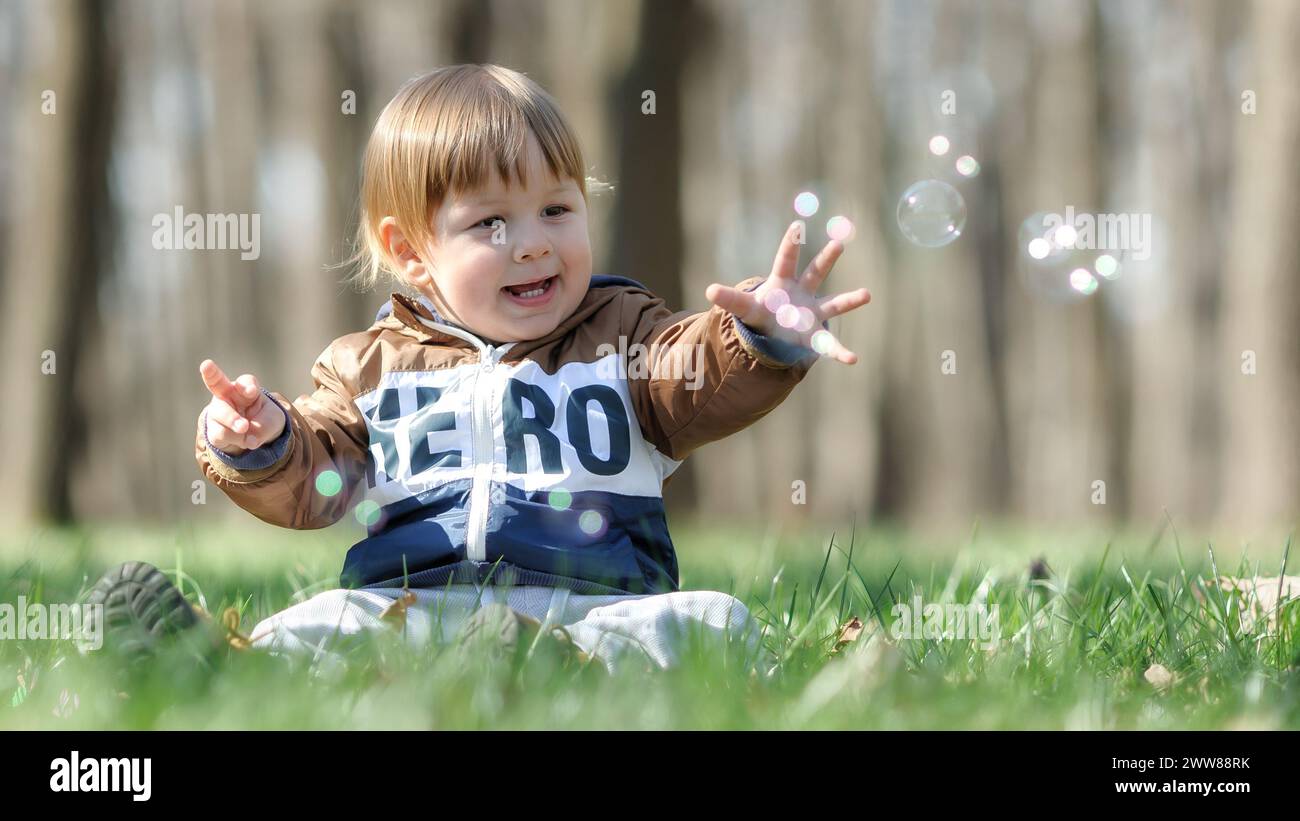 Felice, simpatico, sorridente, bambino seduto su erba verde all'aperto, che gioca con le bolle di sapone. Concetto di amore, cura, genitorialità Foto Stock