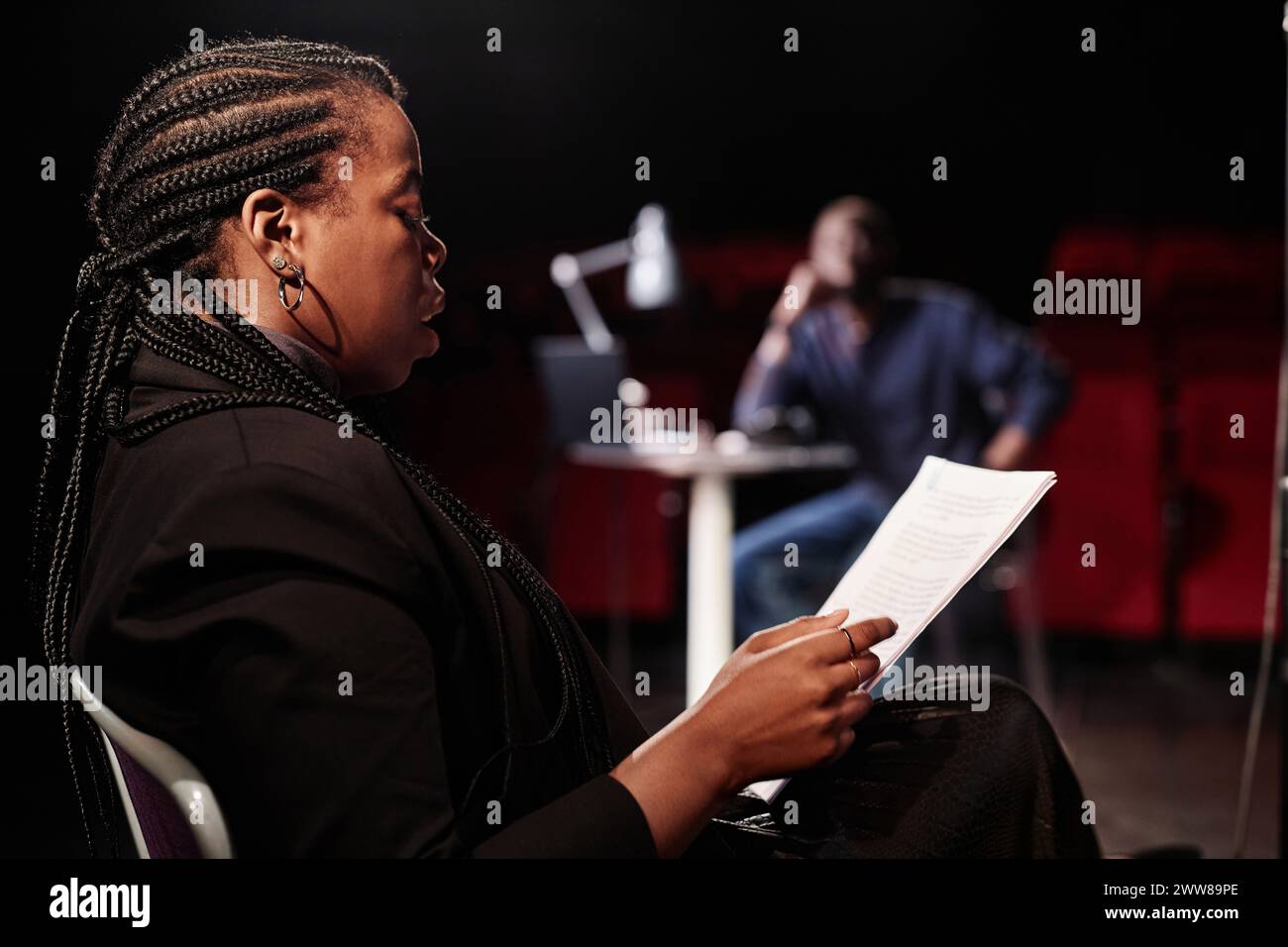 Vista laterale del ritratto di una donna afroamericana che legge la sceneggiatura seduto su una sedia sul palco con spazio fotocopie a bassa illuminazione Foto Stock