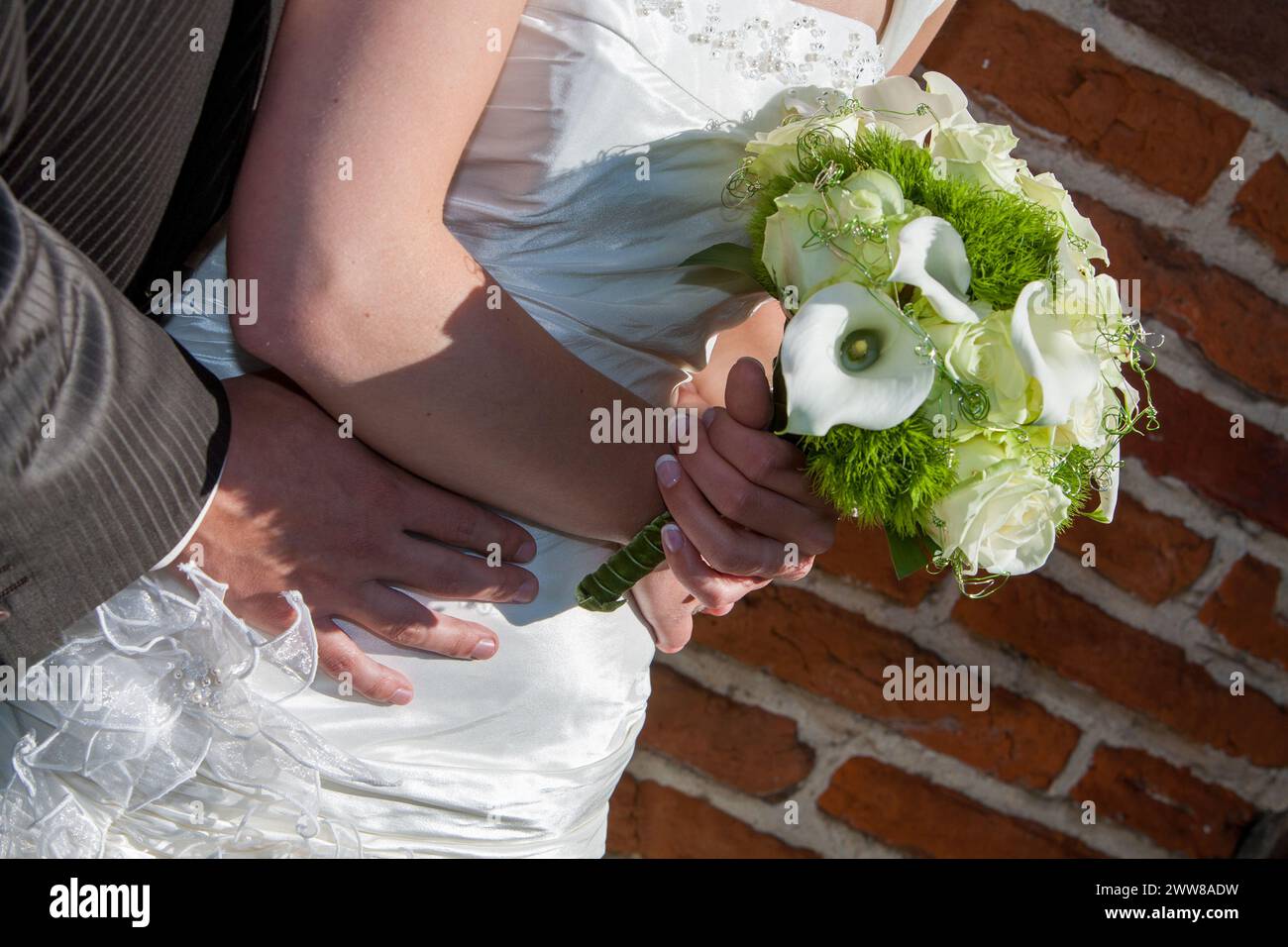 L'immagine incornicia splendidamente le mani agitate di una sposa e di uno sposo sullo sfondo del suo elegante bouquet nuziale e della texture senza tempo di una parete in mattoni, catturando un momento di unità e impegno nella luce dorata del sole. Abbraccio delle promesse: Gli sposi si stringono la mano con il bouquet nuziale. Foto di alta qualità Foto Stock