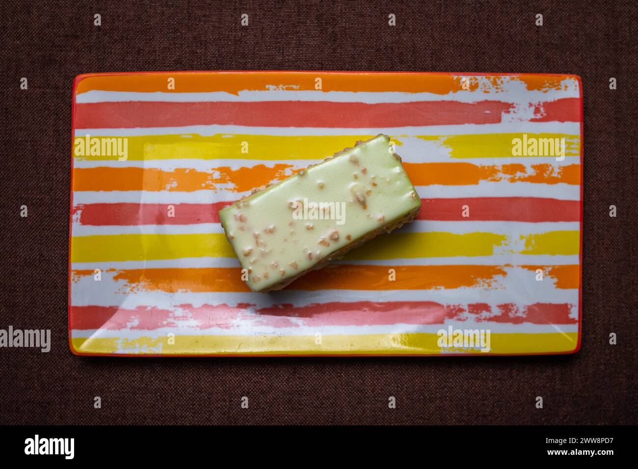 Torta quadrata con brillante glassa bianca su un piatto colorato, catturata in primo piano. La torta è isolata su uno sfondo marrone. Foto Stock