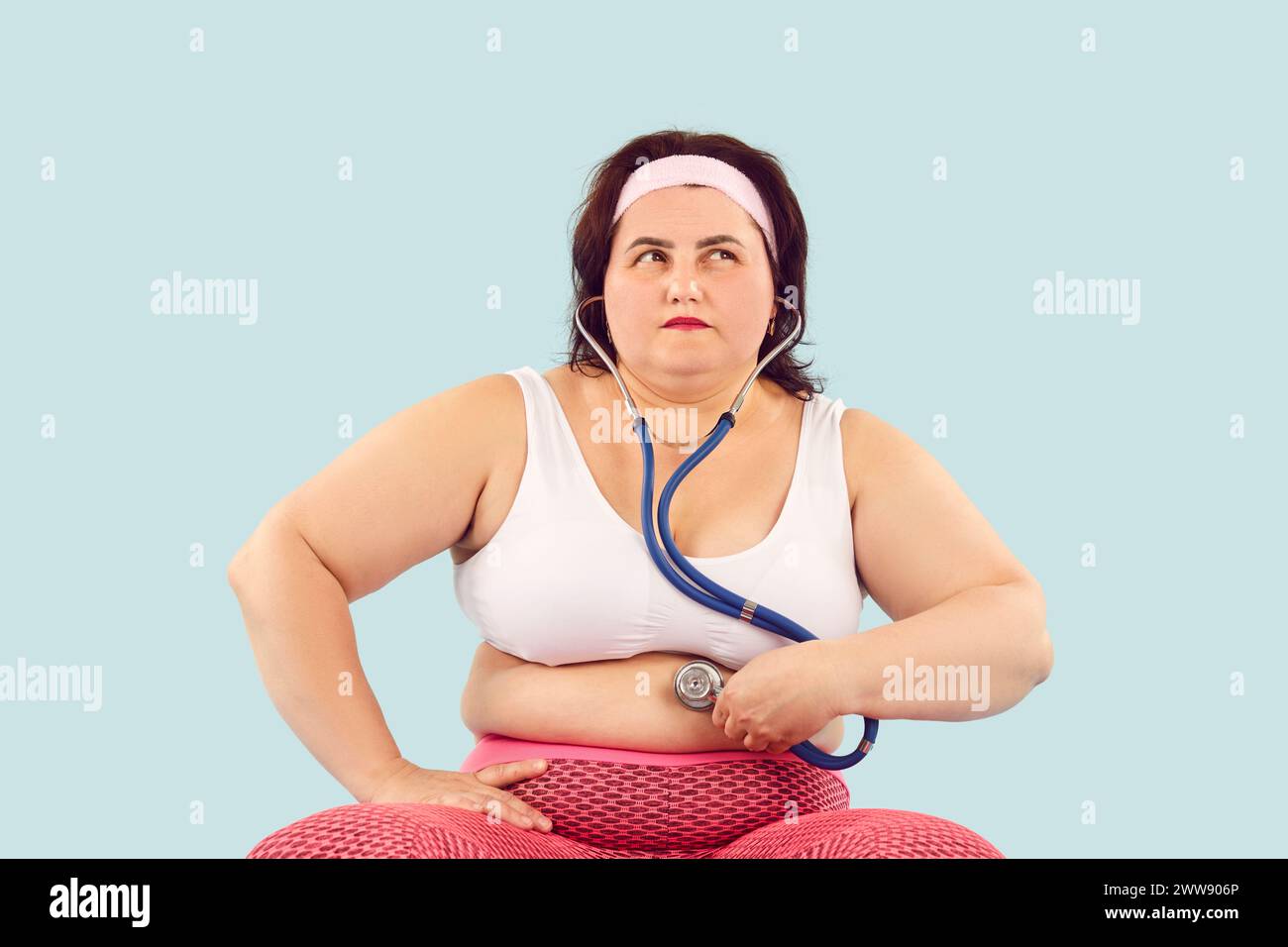 La giovane donna con problemi di obesità e digestione tiene lo stetoscopio sul suo stomaco grasso Foto Stock