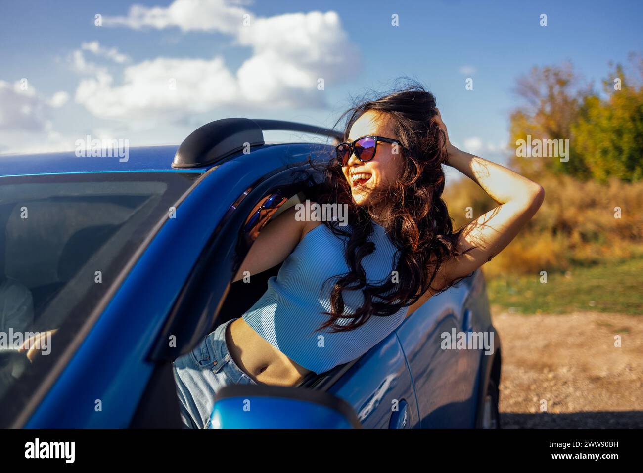 Una giovane asiatica si sporge dal finestrino dell'auto e ride. Una donna coreana sorridente viaggia e si diverte in viaggio. Una ragazza attraente si diverte a guidare e a Foto Stock