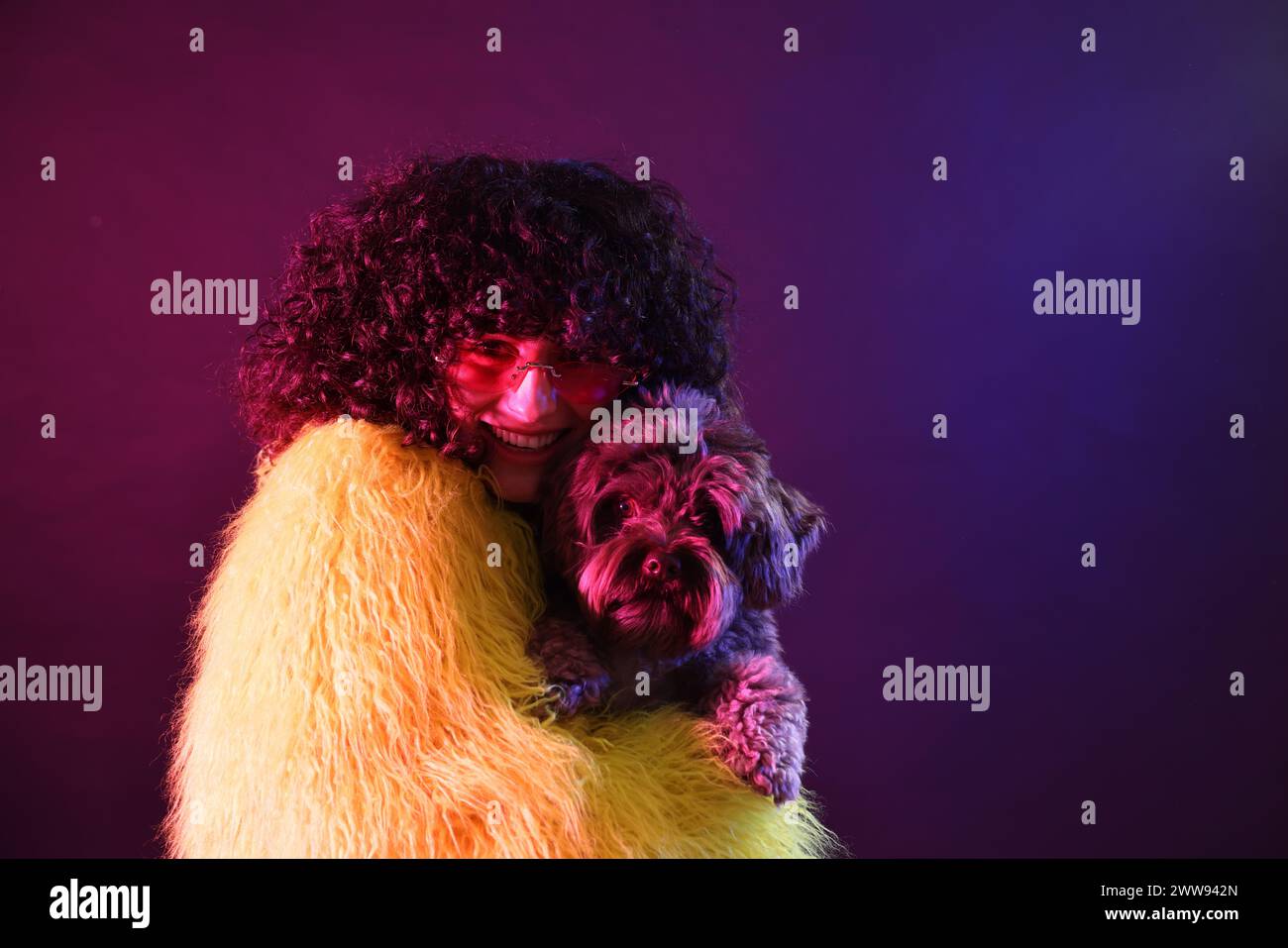 Bella giovane donna con cappotto di pelliccia gialla e occhiali da sole con simpatico cane su sfondo colorato con luci al neon. Spazio per il testo Foto Stock