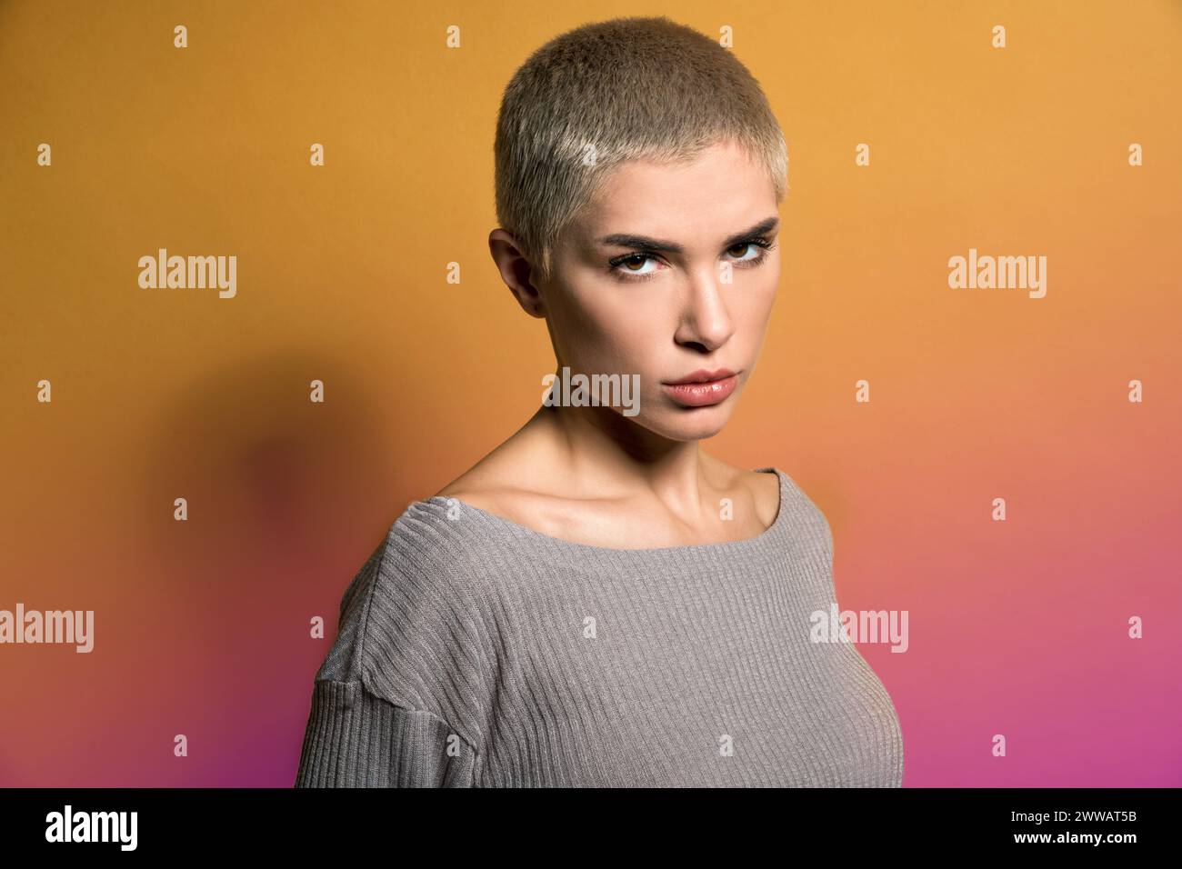 Giovane modella seria con capelli biondi corti in abiti casual che guarda la fotocamera con attenzione su sfondo bicolore sfumato Foto Stock