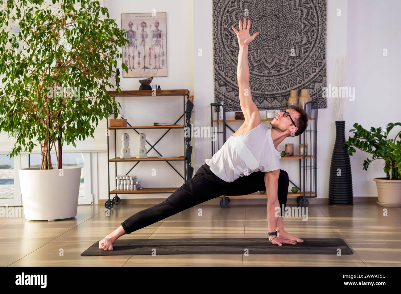 Corpo completo di ragazzo a piedi nudi in abbigliamento sportivo e occhiali da vista che praticano il lunge a rotazione posa yoga sul tappetino durante la sessione di pratica in studio moderno con po Foto Stock