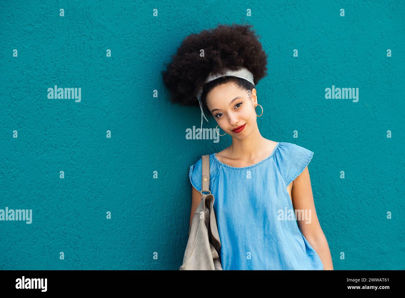 Ritratto di una giovane donna marocchina sorridente in trucco con fascia in capelli afro che guarda la fotocamera mentre sta in piedi con la borsa su sfondo blu in d Foto Stock