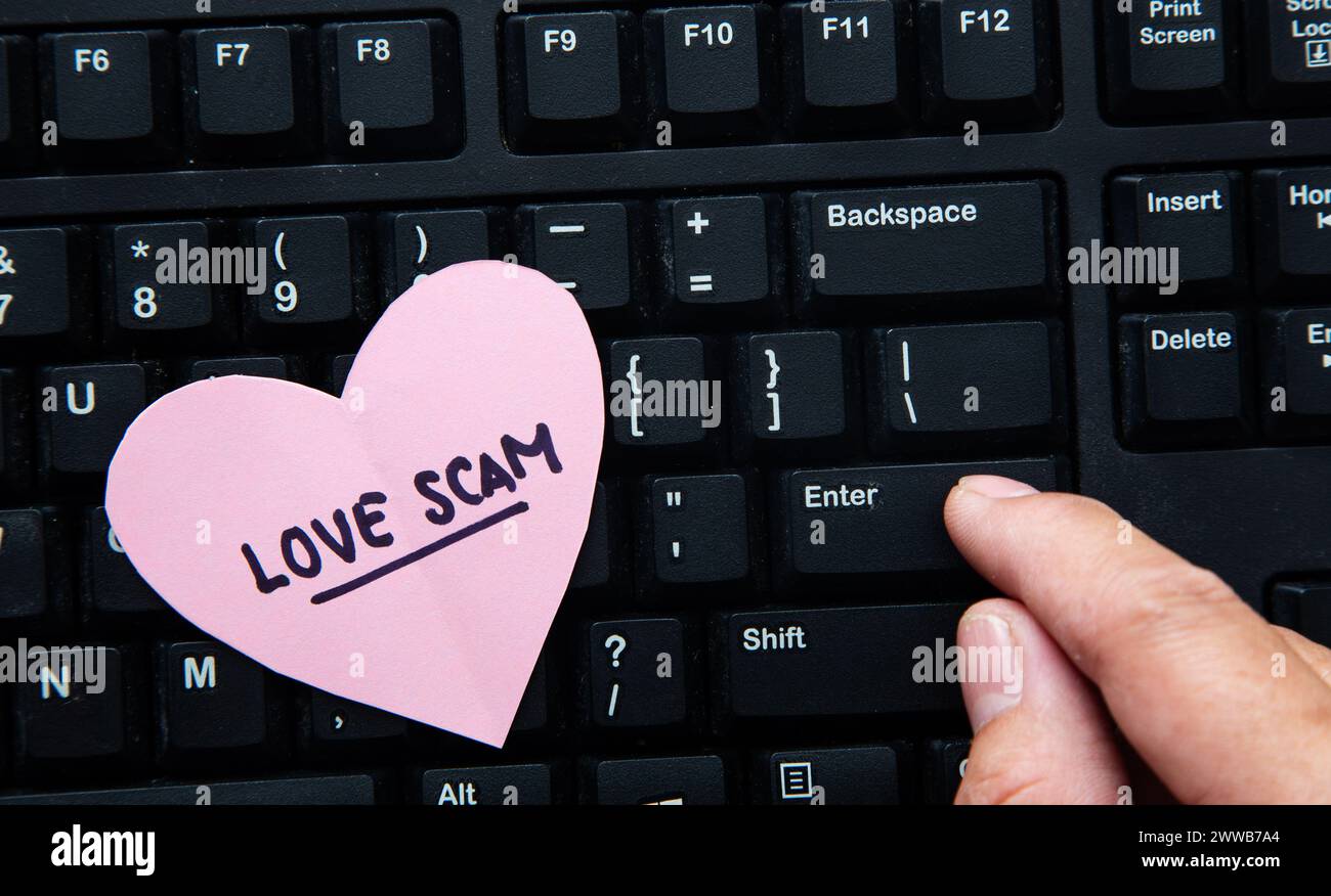 Cuore rosa con testo di truffa amorosa posizionato su una tastiera del computer mentre un dito preme il tasto Invio Foto Stock