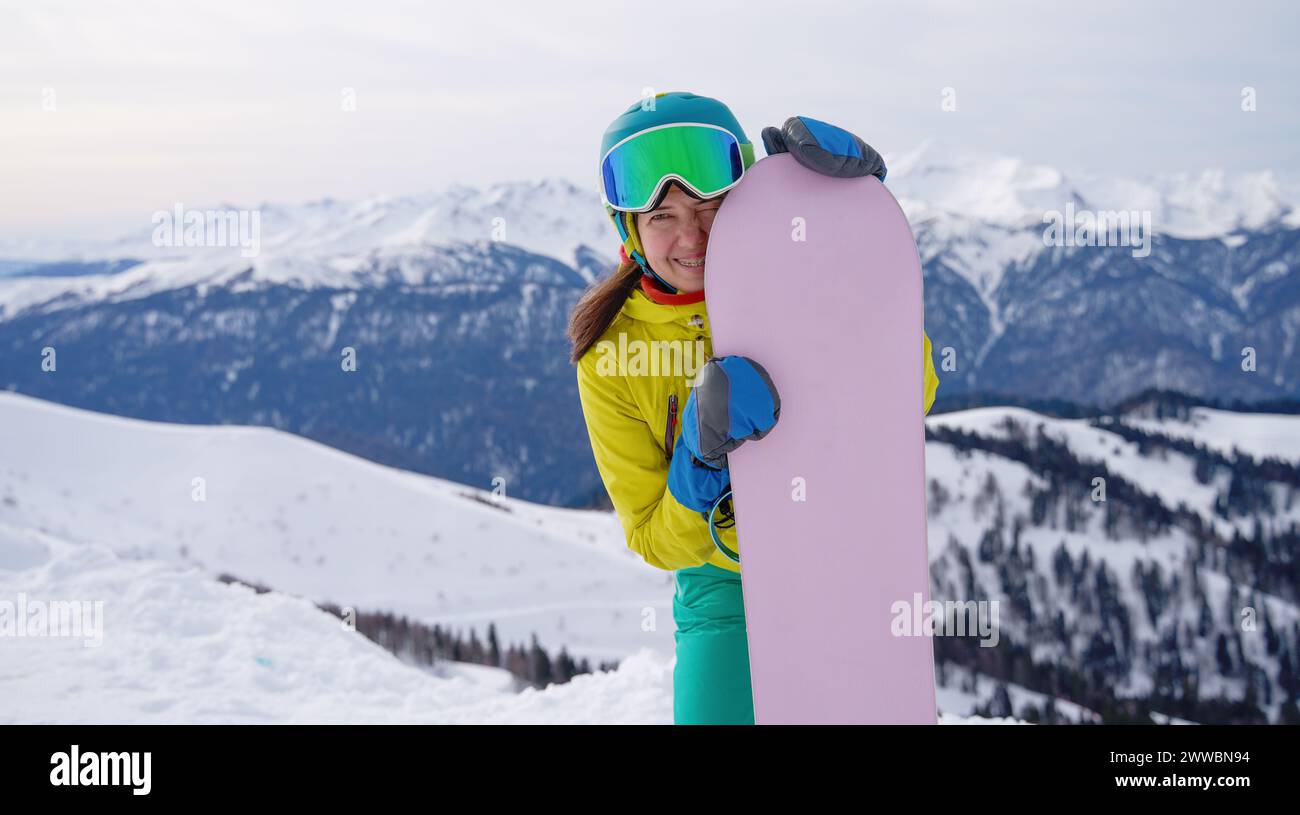 Un giovane snowboarder abbraccia la tavola, sorridendo contro la luce calante del cielo crepuscolo sulle montagne innevate, pronto per le emozioni del giorno successivo. Foto Stock