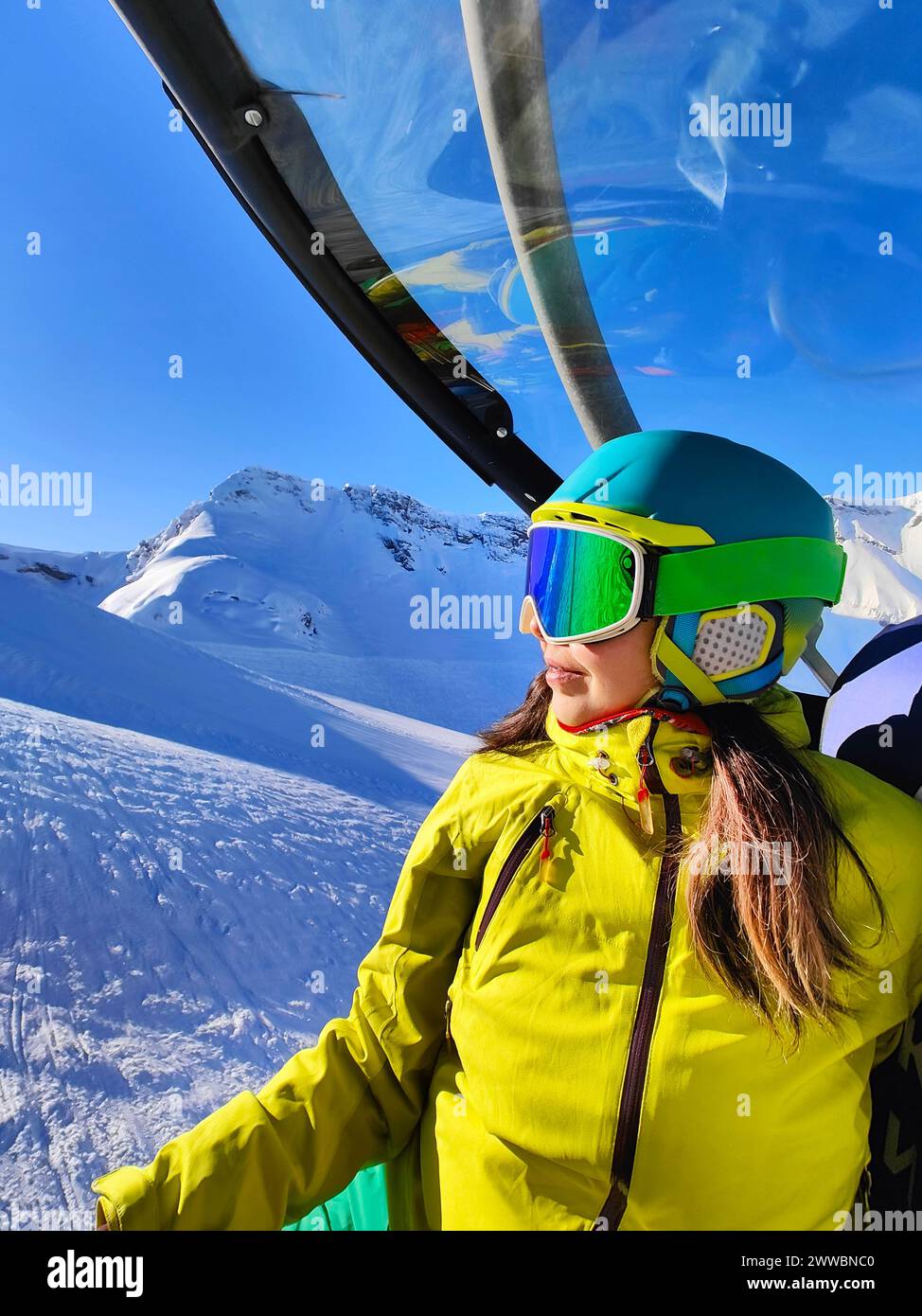 Uno sciatore guarda fuori da una funivia, il cielo azzurro e le cime innevate che si riflettono negli occhiali, catturando l'attesa delle piste. Foto Stock