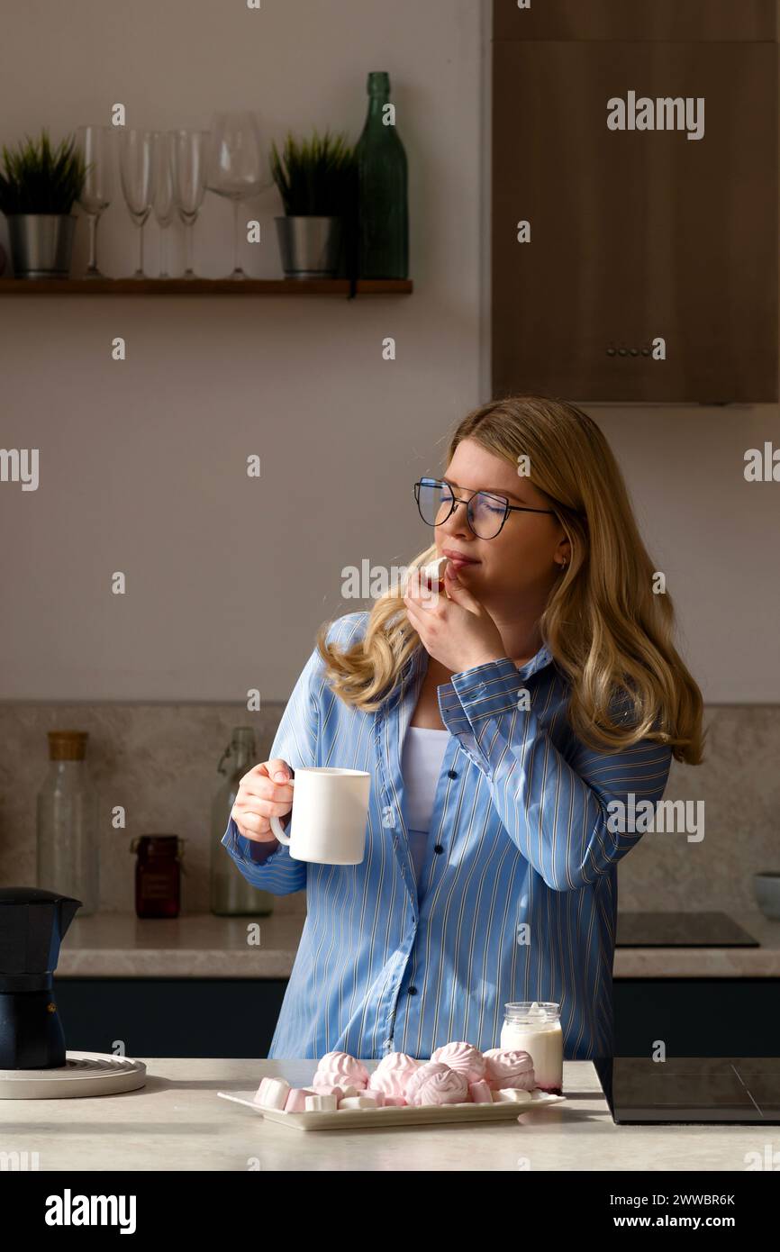 Donna che assaggia il caffè, cucina tranquilla. Una ragazza con camicia a righe blu ama i marshmallow sorseggiando un caffè mattutino Foto Stock