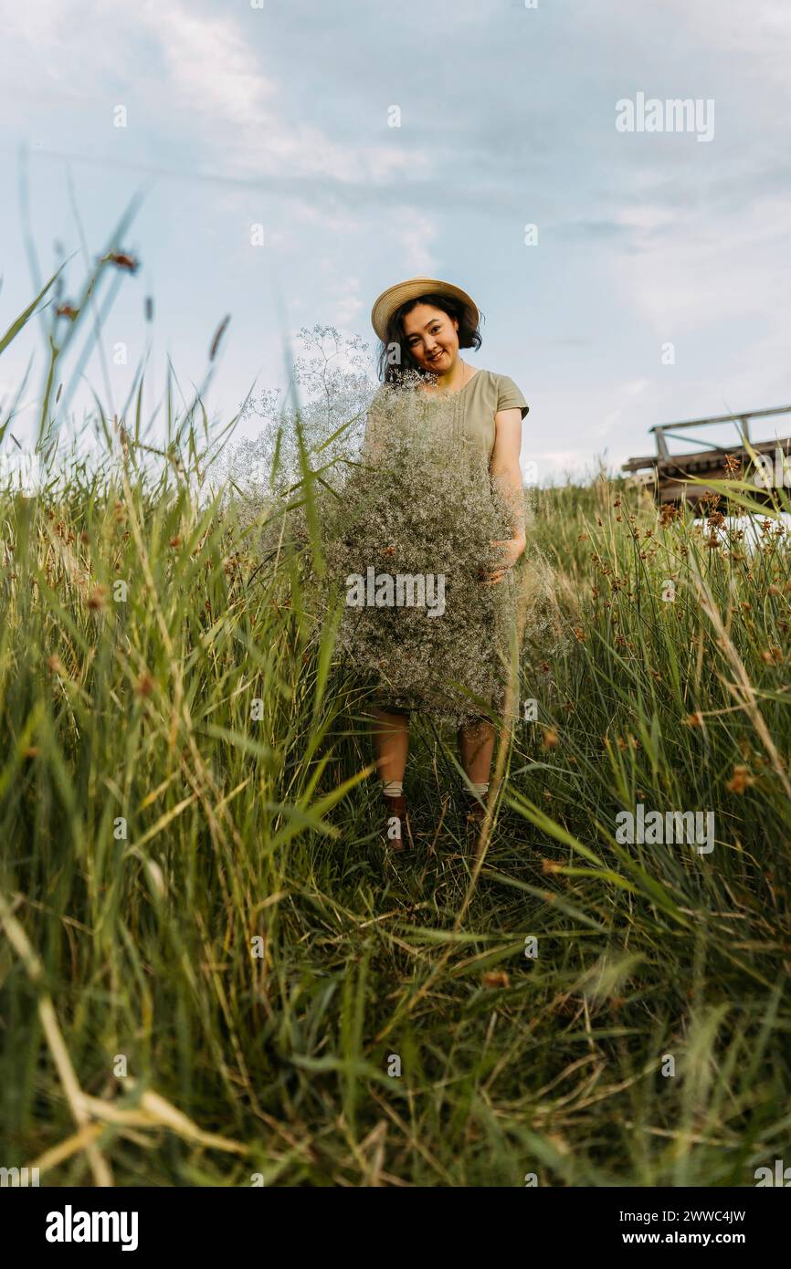 Donna matura sorridente che tiene un mazzo di fiori di gypsophila in mezzo all'erba nel campo Foto Stock