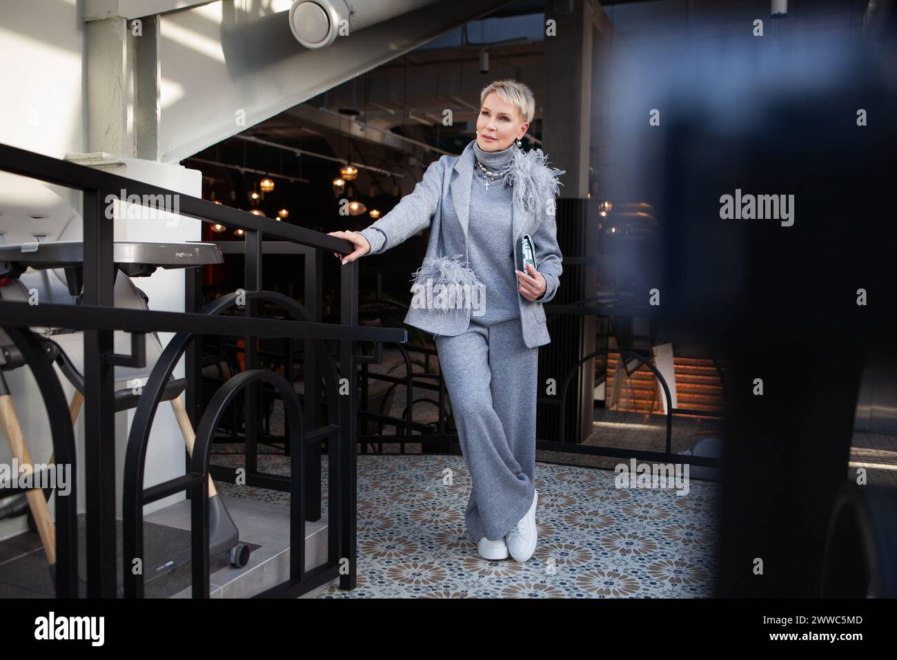 Donna matura attraente ed elegante in pantalone grigio con giacca e dolcevita, accessoriata con borsa e scarpe bianche. Il look perfetto per succ Foto Stock