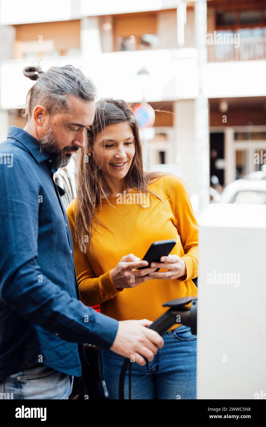 Coppia che paga tramite smartphone alla stazione di ricarica per veicoli elettrici Foto Stock