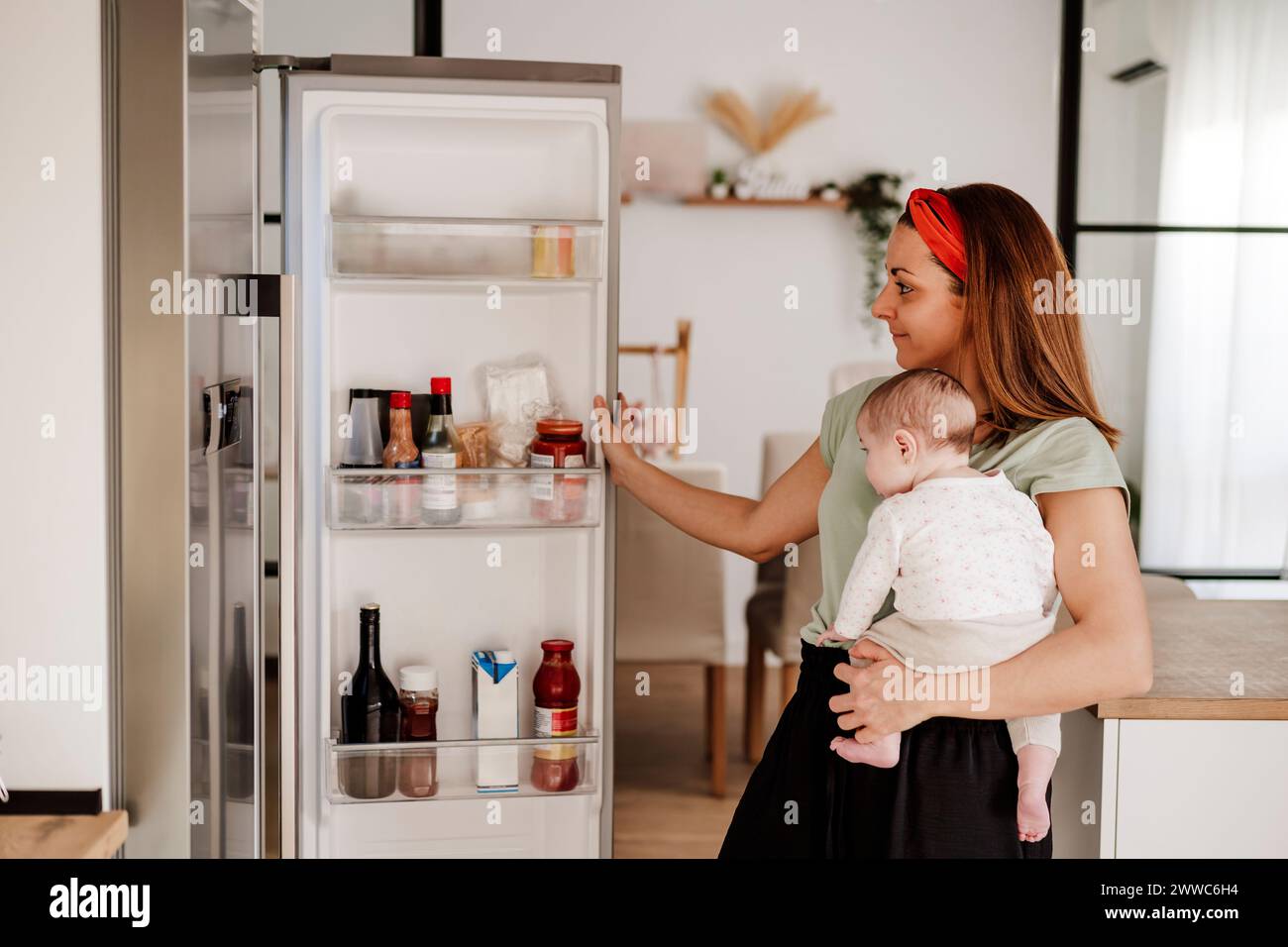 Donna sorridente che porta la bambina e apre la porta del frigorifero Foto Stock
