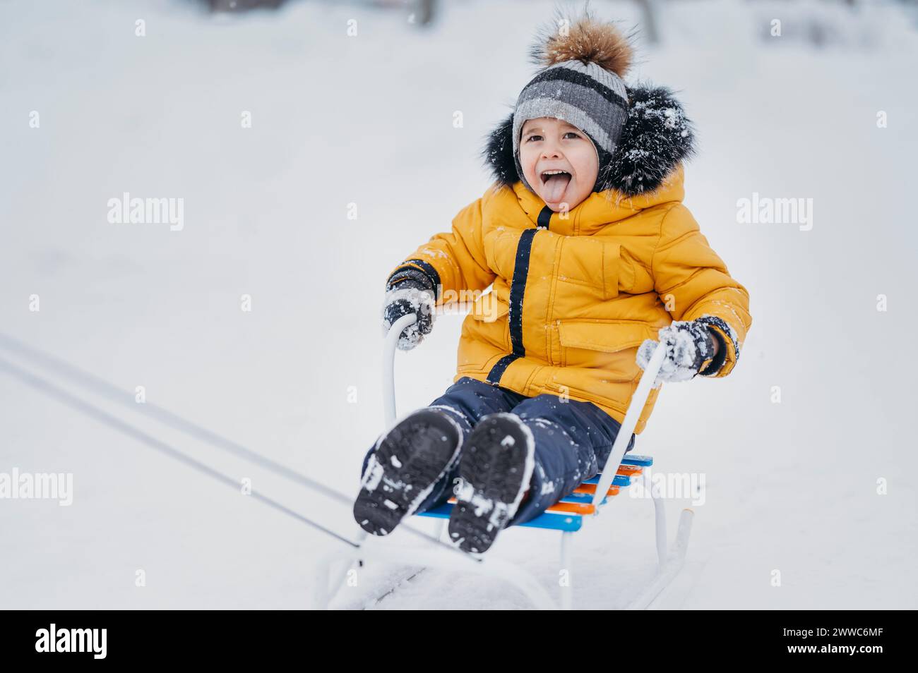 Un ragazzo allegro che indossa abiti caldi e si diverte a fare un giro in slitta sulla neve in inverno Foto Stock