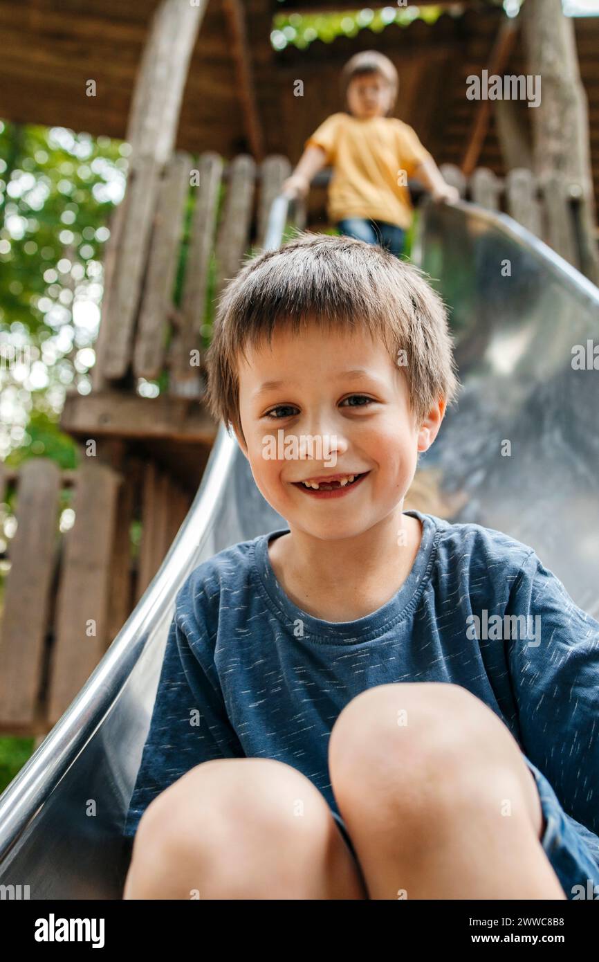 Il ragazzo felice scivola giù sullo scivolo con il fratello sullo sfondo al parco giochi Foto Stock