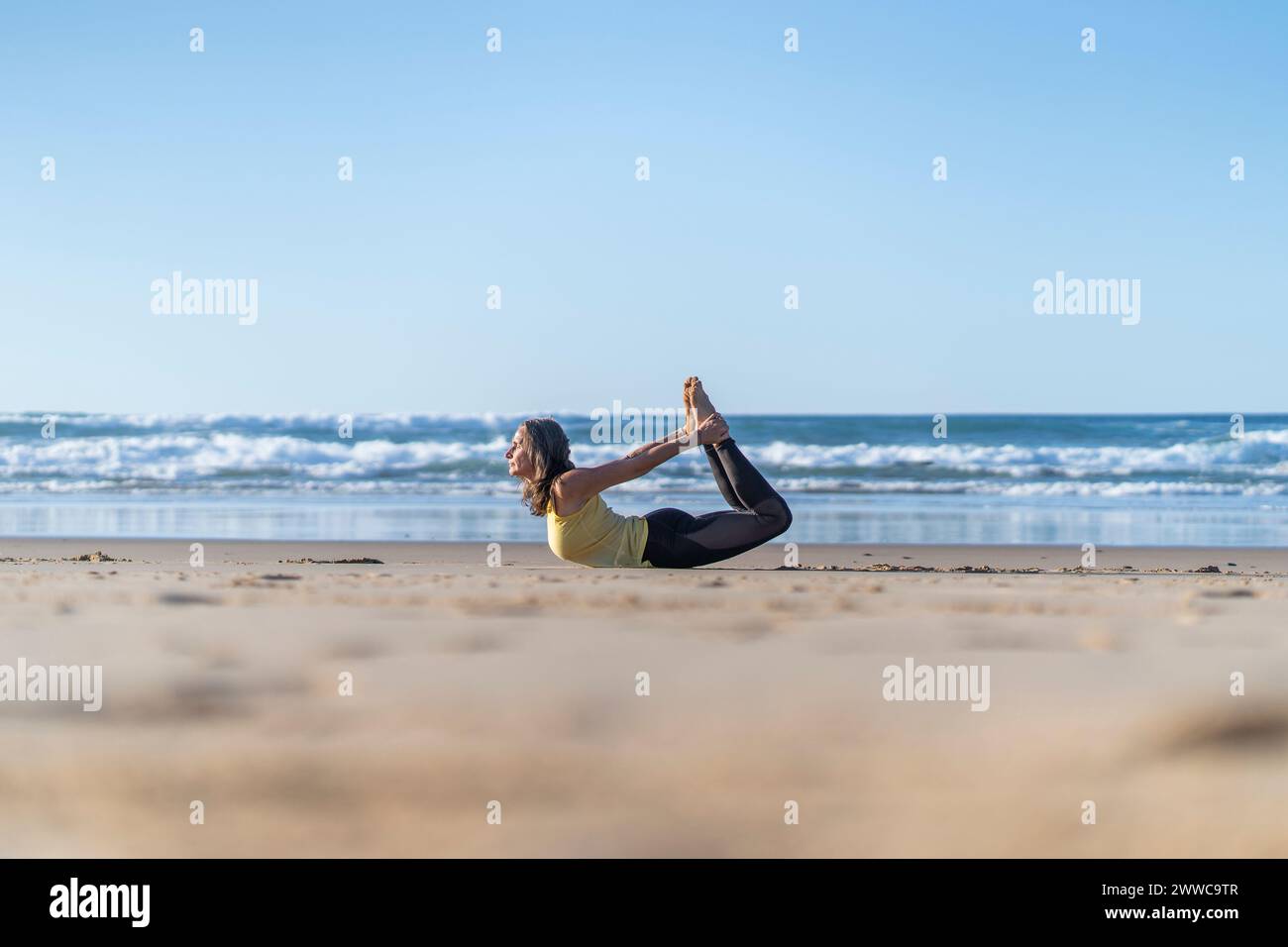 Donna flessibile e matura che pratica la posa dello yoga di prua sulla spiaggia Foto Stock