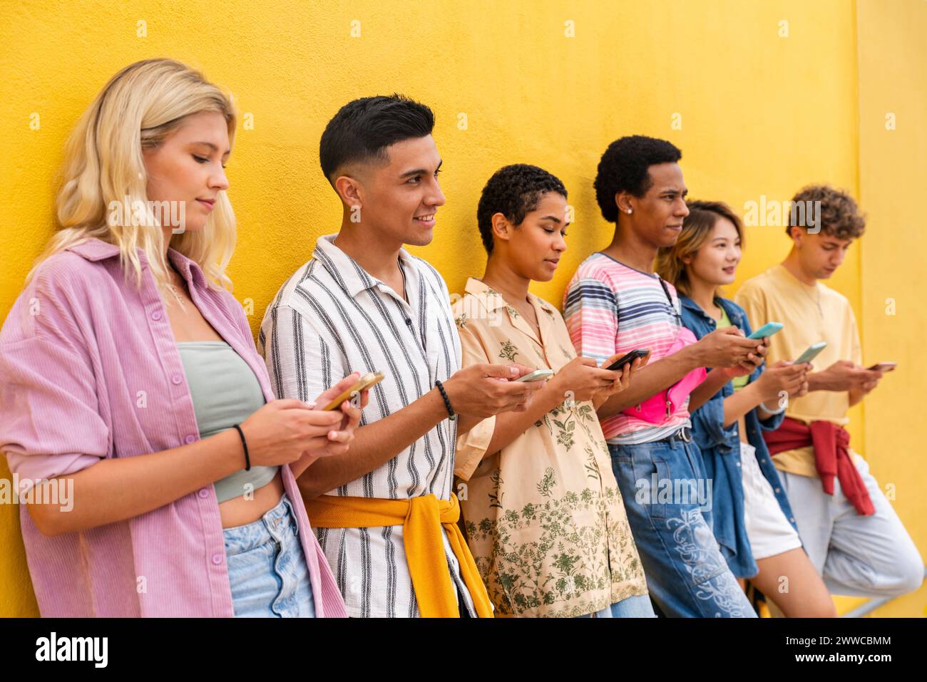 Gruppo di amici appoggiati contro il muro giallo utilizzando i loro smartphone Foto Stock