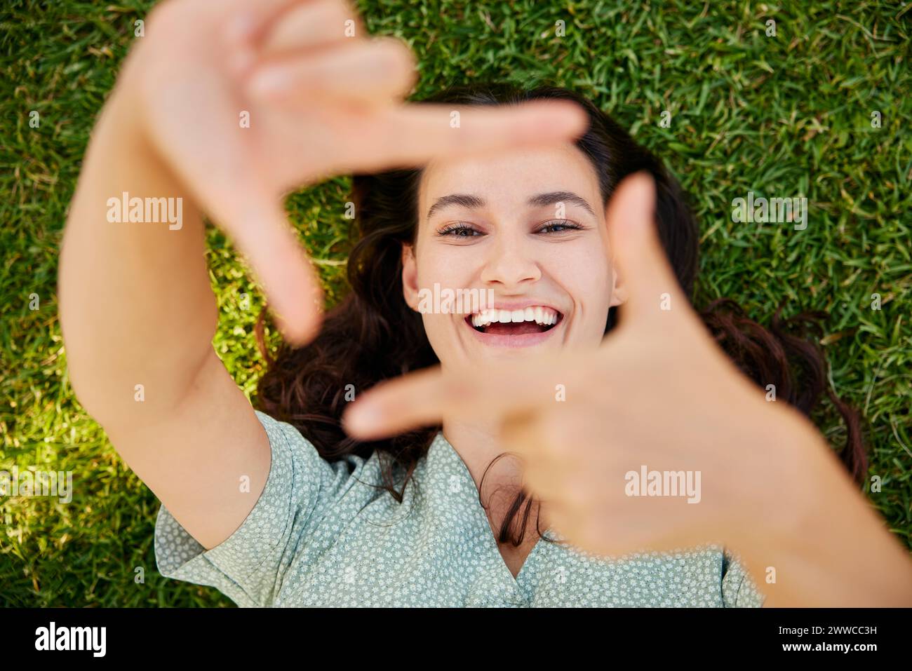 Donna felice che guarda attraverso la cornice delle dita e sdraiata sull'erba Foto Stock