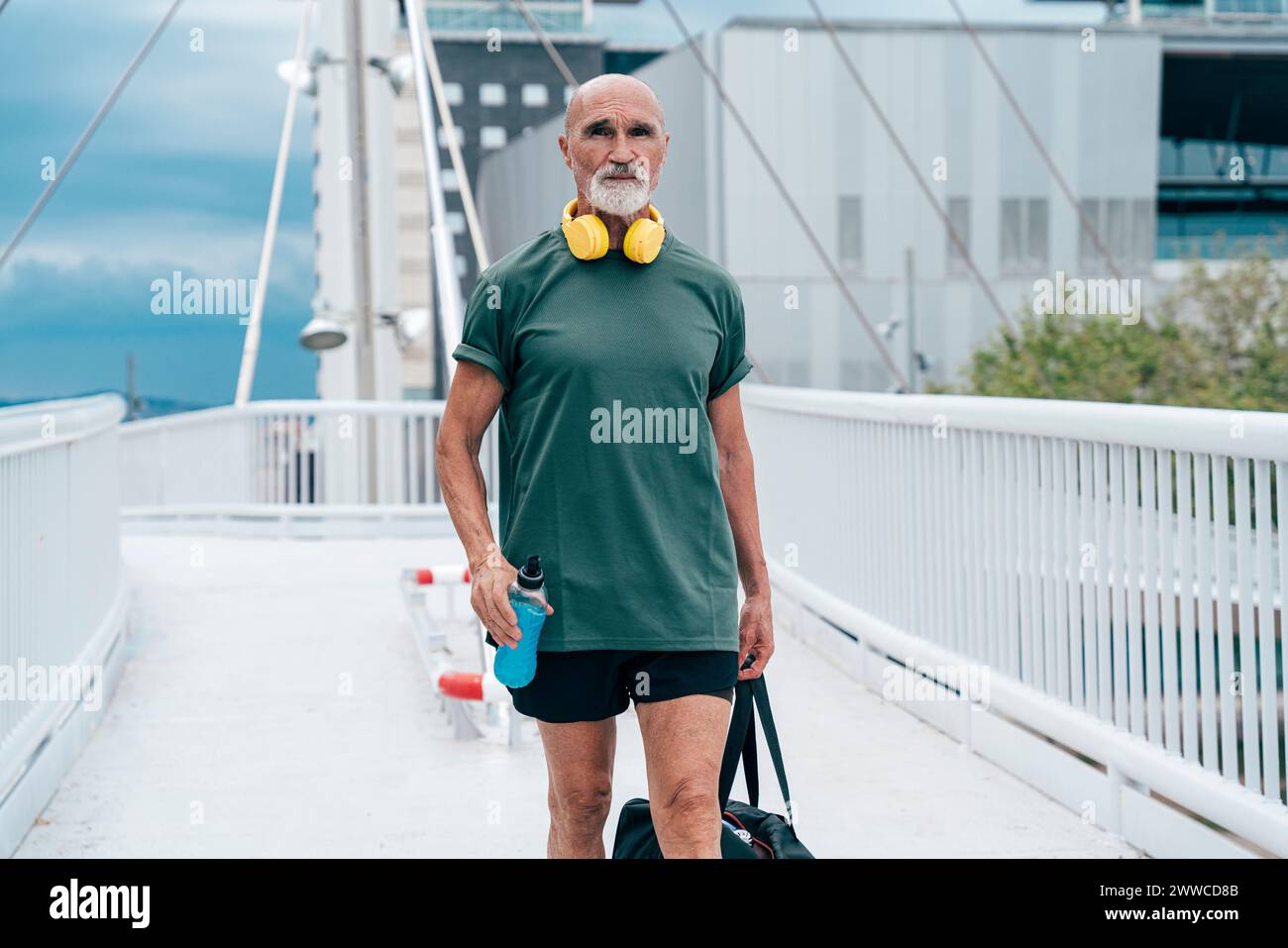 Anziano in pensione che cammina con una bottiglia di bevanda energetica e una borsa sul ponte pedonale Foto Stock