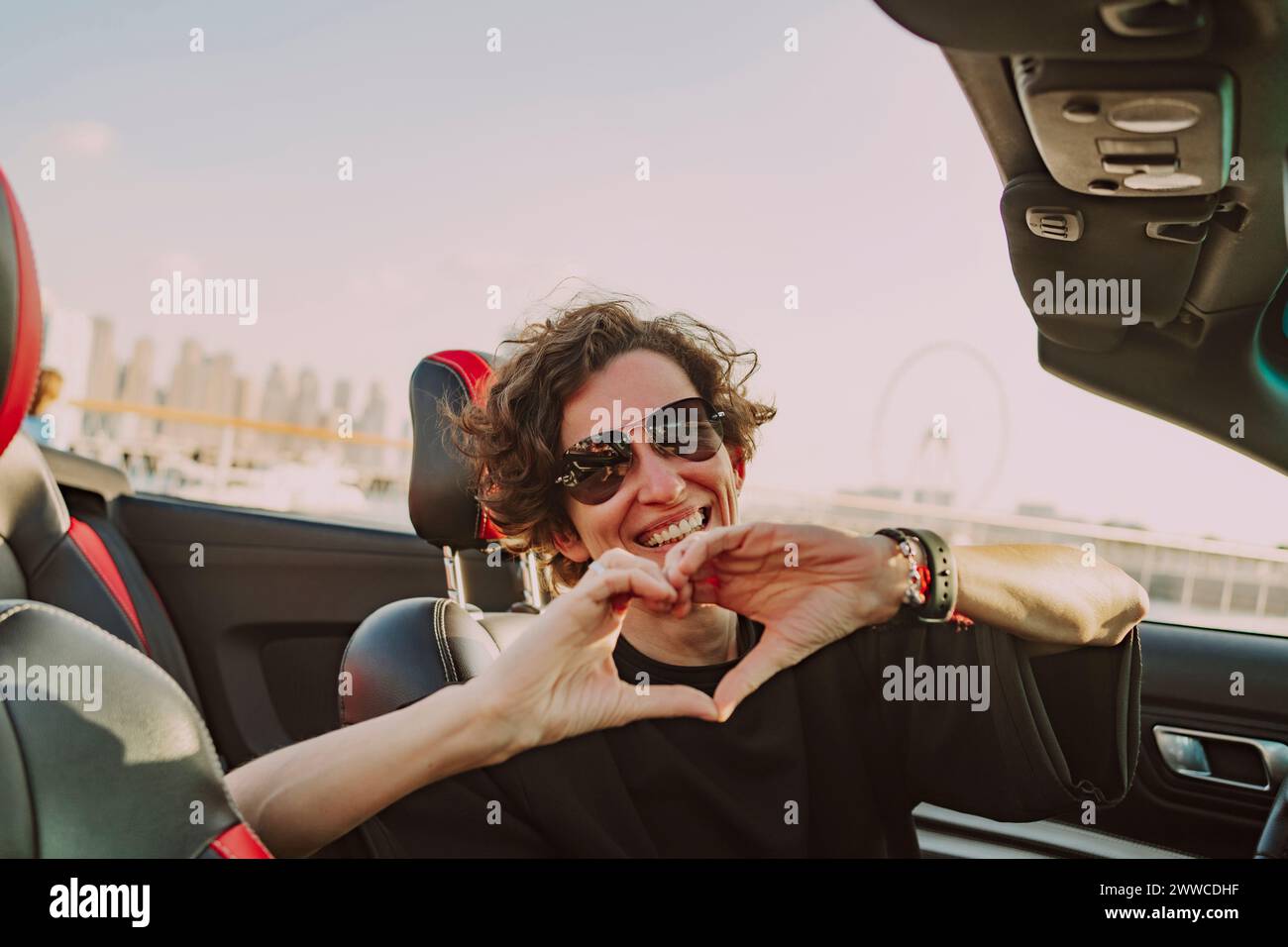 Donna felice che indossa occhiali da sole che mostrano la forma del cuore seduto in una vettura convertibile nelle giornate di sole Foto Stock