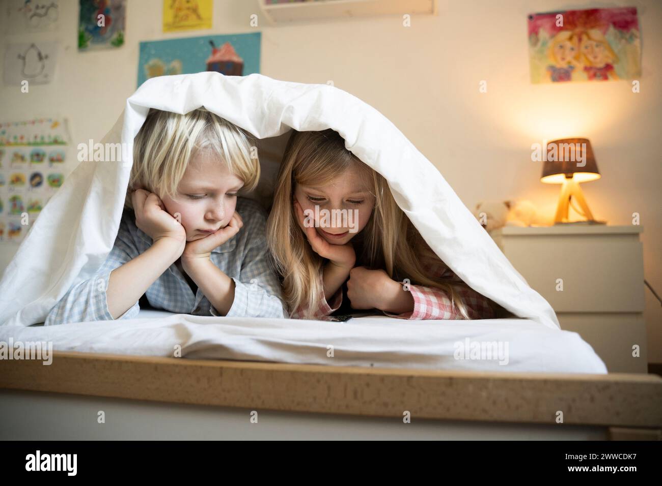 Fratelli che guardano lo smartphone sdraiati sotto la coperta sul letto di casa Foto Stock