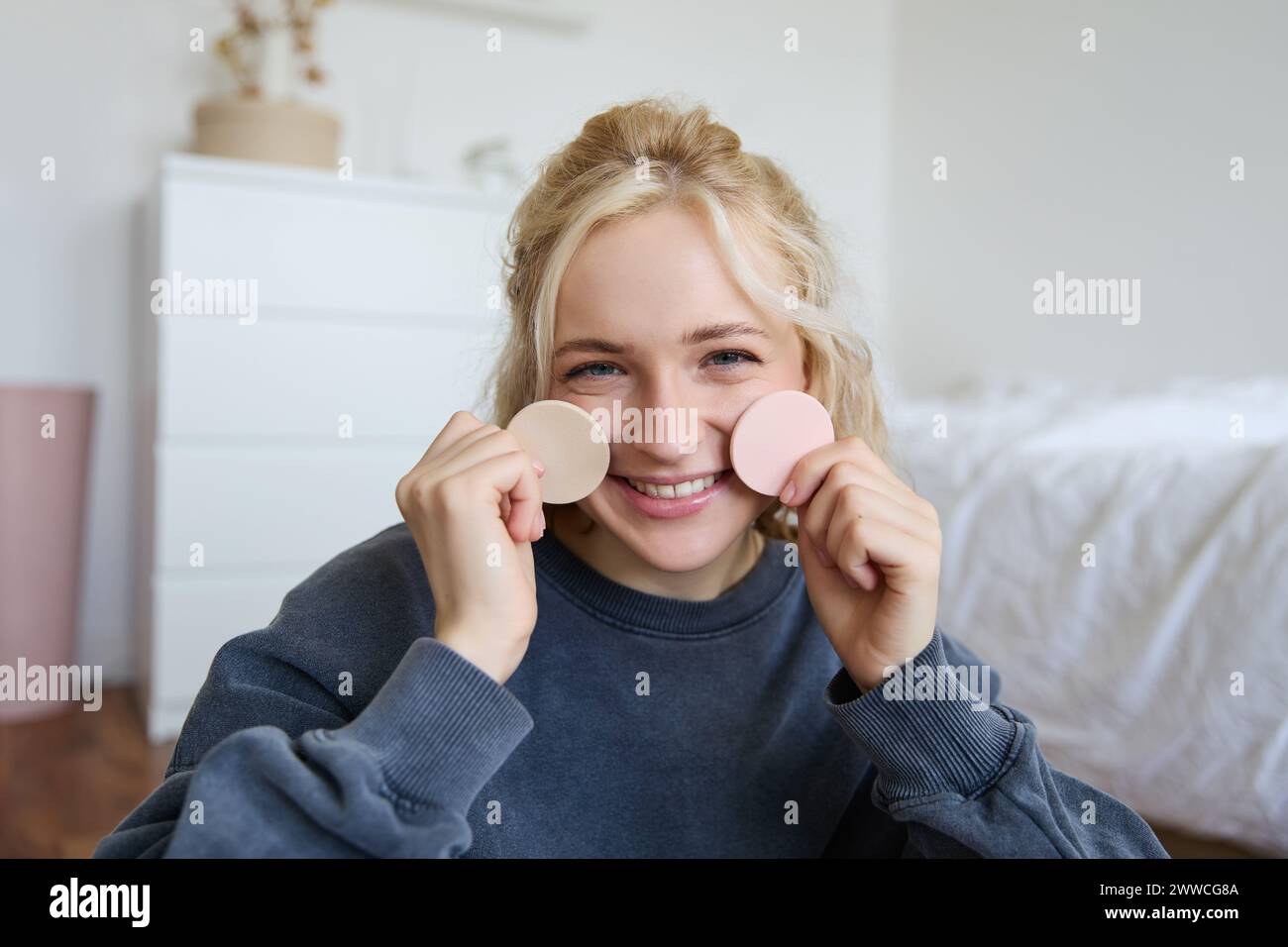 Primo piano di una giovane donna felice e bella, che mostra prodotti di bellezza sulla fotocamera, consiglia il trucco, seduto sul pavimento in camera da letto Foto Stock