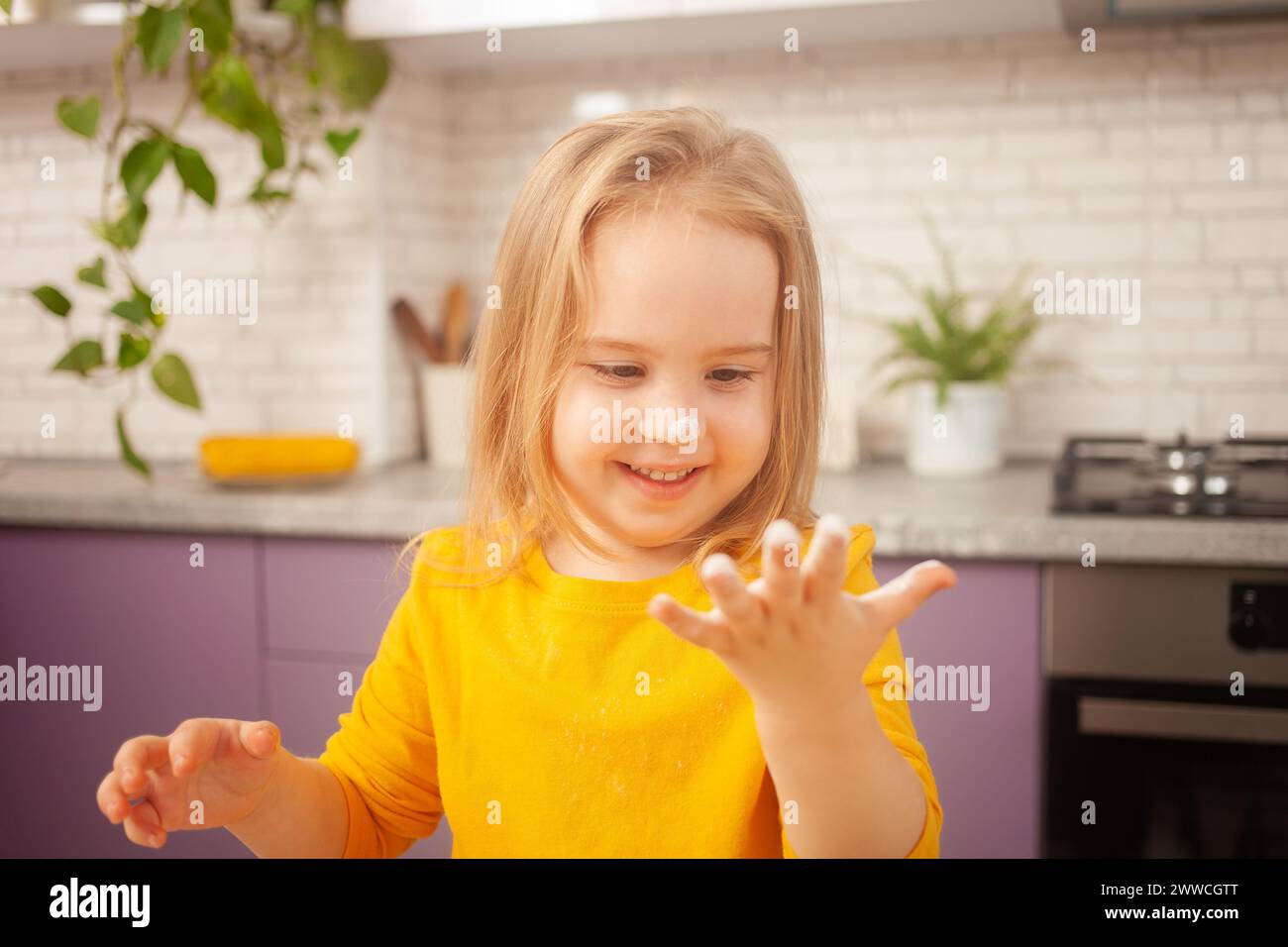la bambina di tre anni in giacca gialla si sta preparando a cucinare il cibo in cucina. Ritratto di un bambino felice. mettere a mano la farina Foto Stock