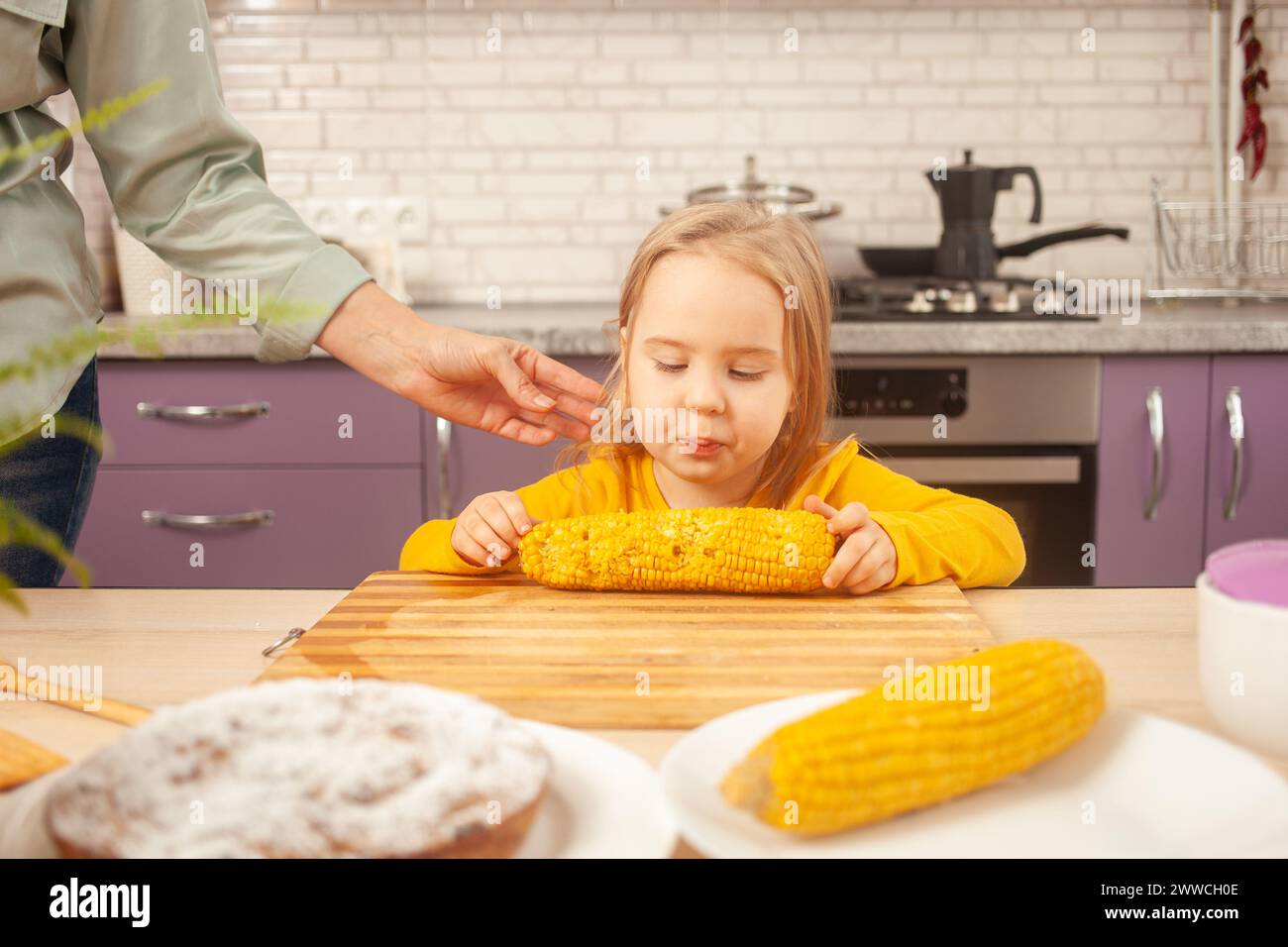il bambino mangia mais dolce bollito nella cucina viola della casa. Ragazza divertente a tavola. Foto Stock
