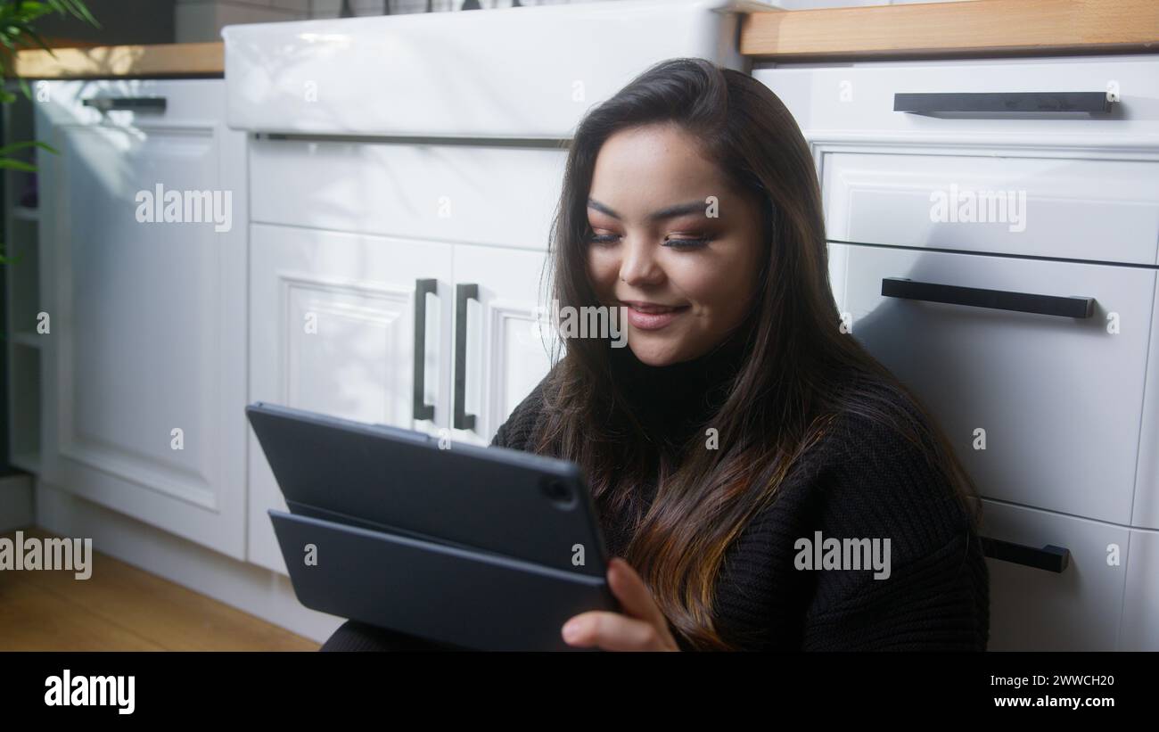 Donna asiatica seduto sul pavimento in cucina, divertendosi con un tablet navigando sul Web, giocando, facendo shopping su Internet, utilizzando app di social media Foto Stock