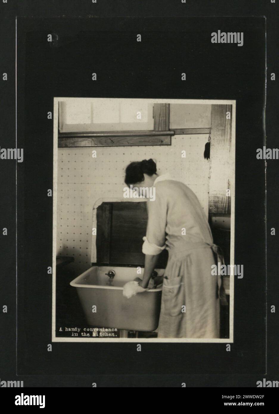 Descrizione: Una comoda comodità in cucina. Luogo: Canada Data: 1920-1929 donna, casa, canada, casa, in piedi, lavello, interni, lavoro, decorazione, lavabo, grembiule, domestico Foto Stock