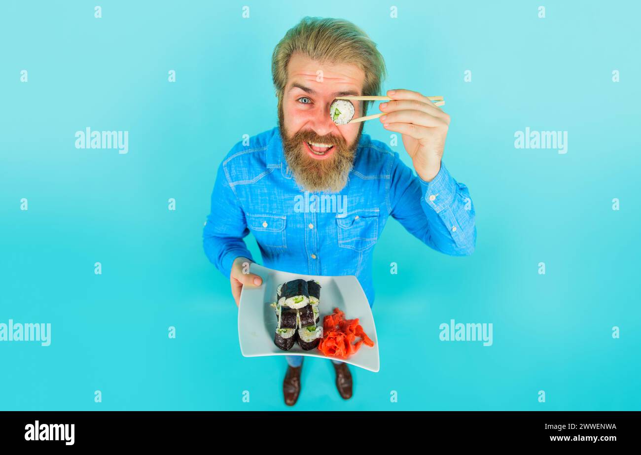 Uomo sorridente con barba in denim casual che copre l'occhio con sushi roll. Cucina tradizionale giapponese. Bell'uomo con un piatto di delizioso maki sushi Foto Stock
