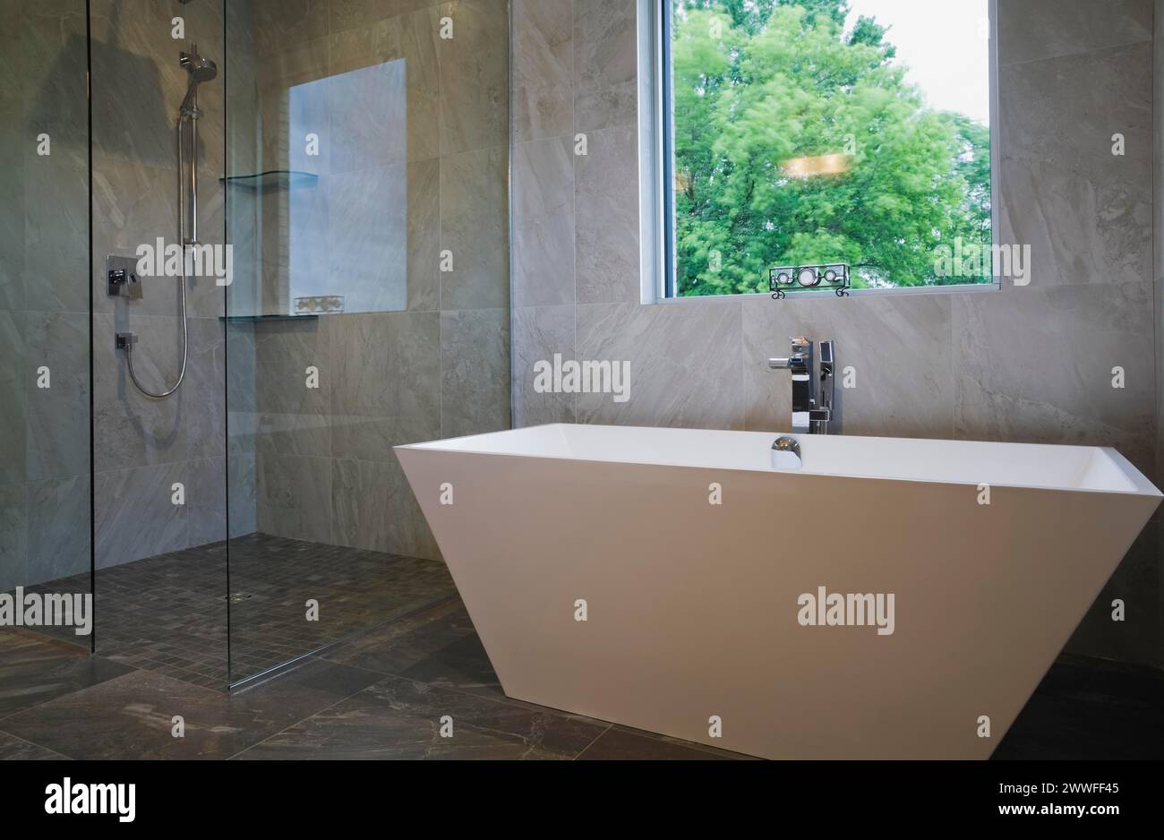 Cabina doccia in vetro trasparente e vasca bianca a forma di vaso indipendente nel bagno con pavimento in piastrelle grigie in ceramica e pareti interne al piano terra Foto Stock