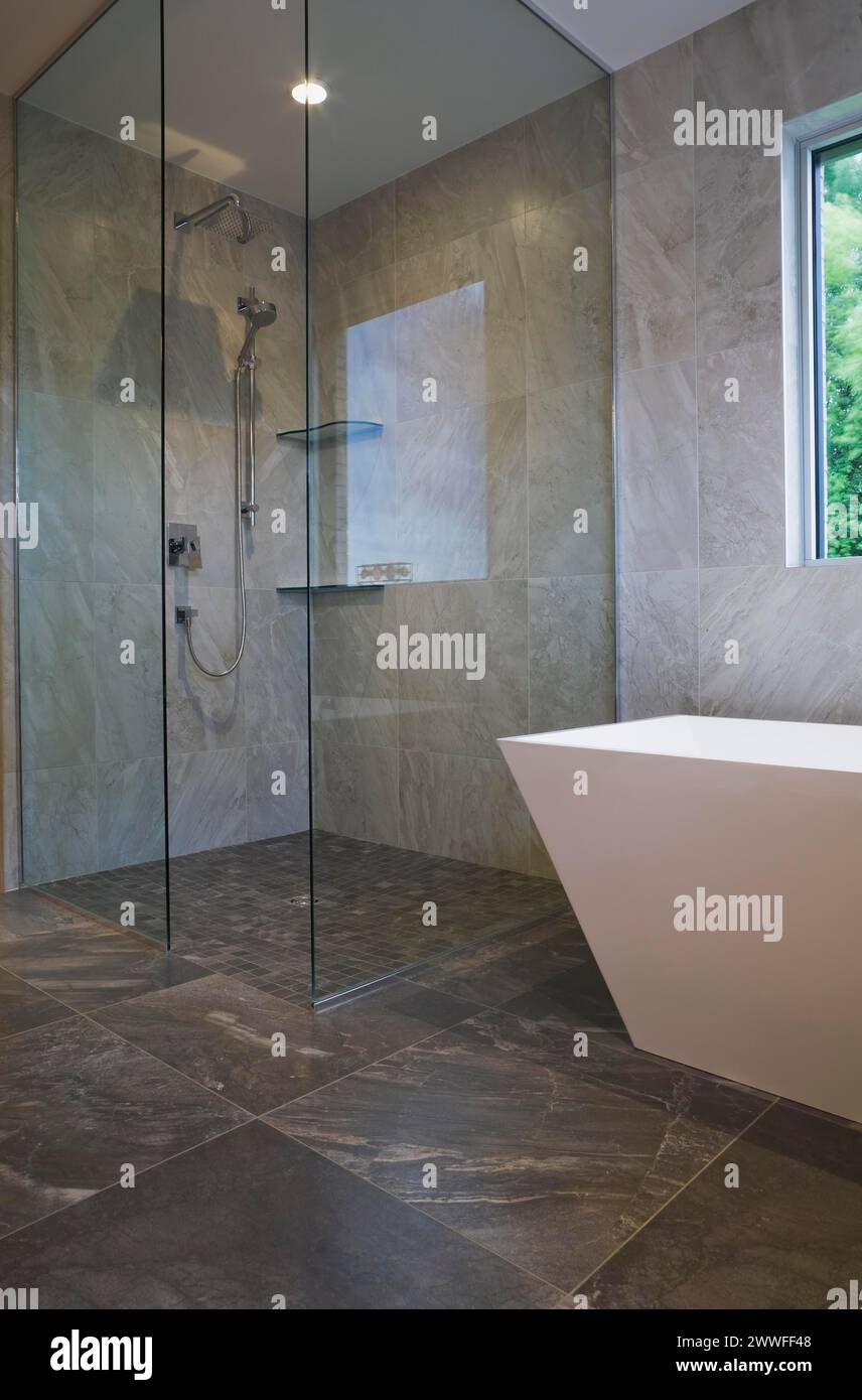 Cabina doccia in vetro trasparente e vasca bianca a forma di vaso indipendente nel bagno con pavimento in piastrelle grigie in ceramica e pareti interne al piano terra Foto Stock