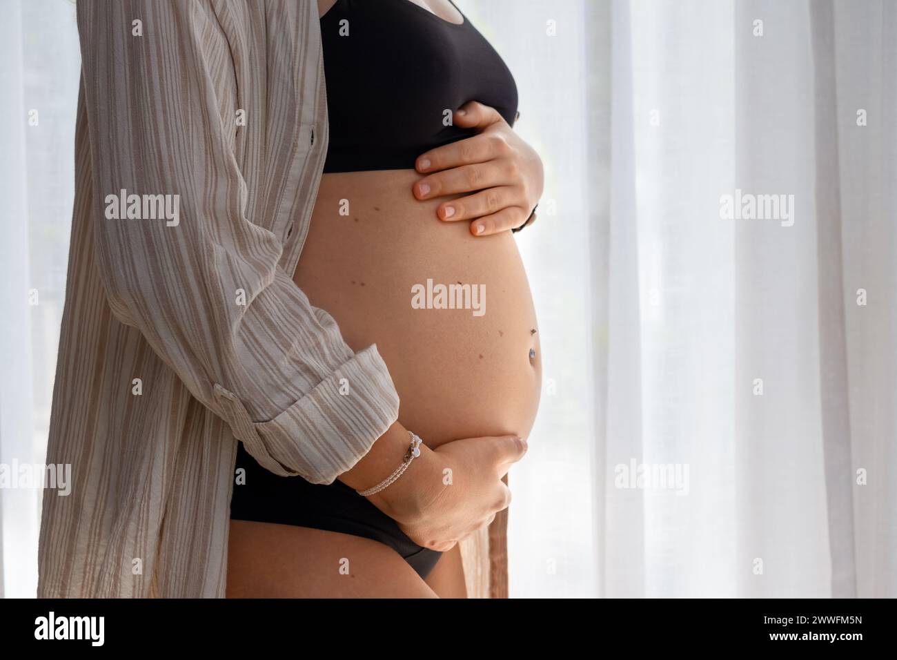 Una ragazza tiene la pancia incinta in una camicia vicino alla finestra Foto Stock