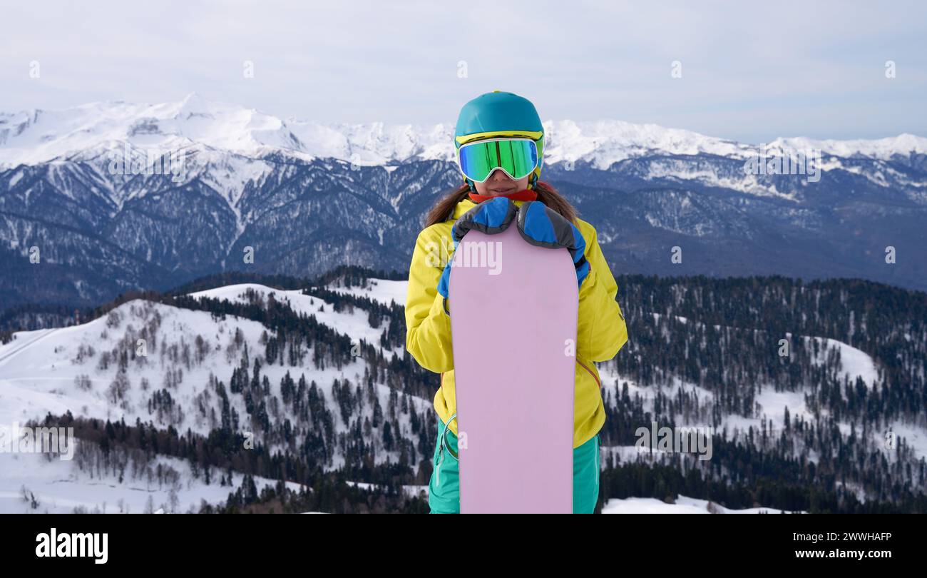 Uno snowboarder con un sorriso avvolgente abbraccia la tavola, davanti a una vista mozzafiato delle vette alpine, incarnando la gioia degli sport invernali. Foto Stock