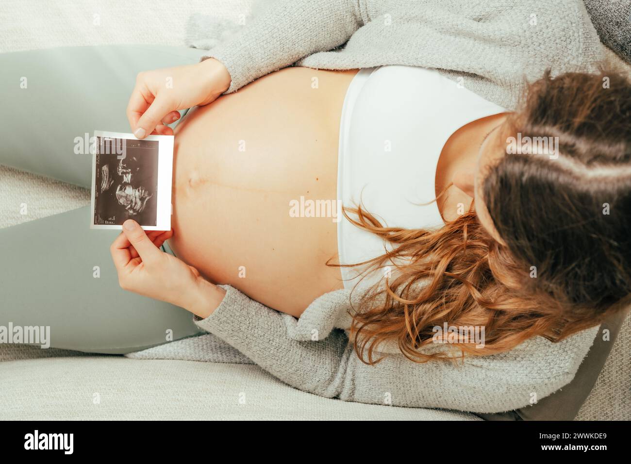 Descrizione: Vista dall'alto di una donna seduta su un divano che guarda un'immagine ecografica del suo bambino nell'ultima fase della gravidanza. Gravidanza, terzo trimestre Foto Stock