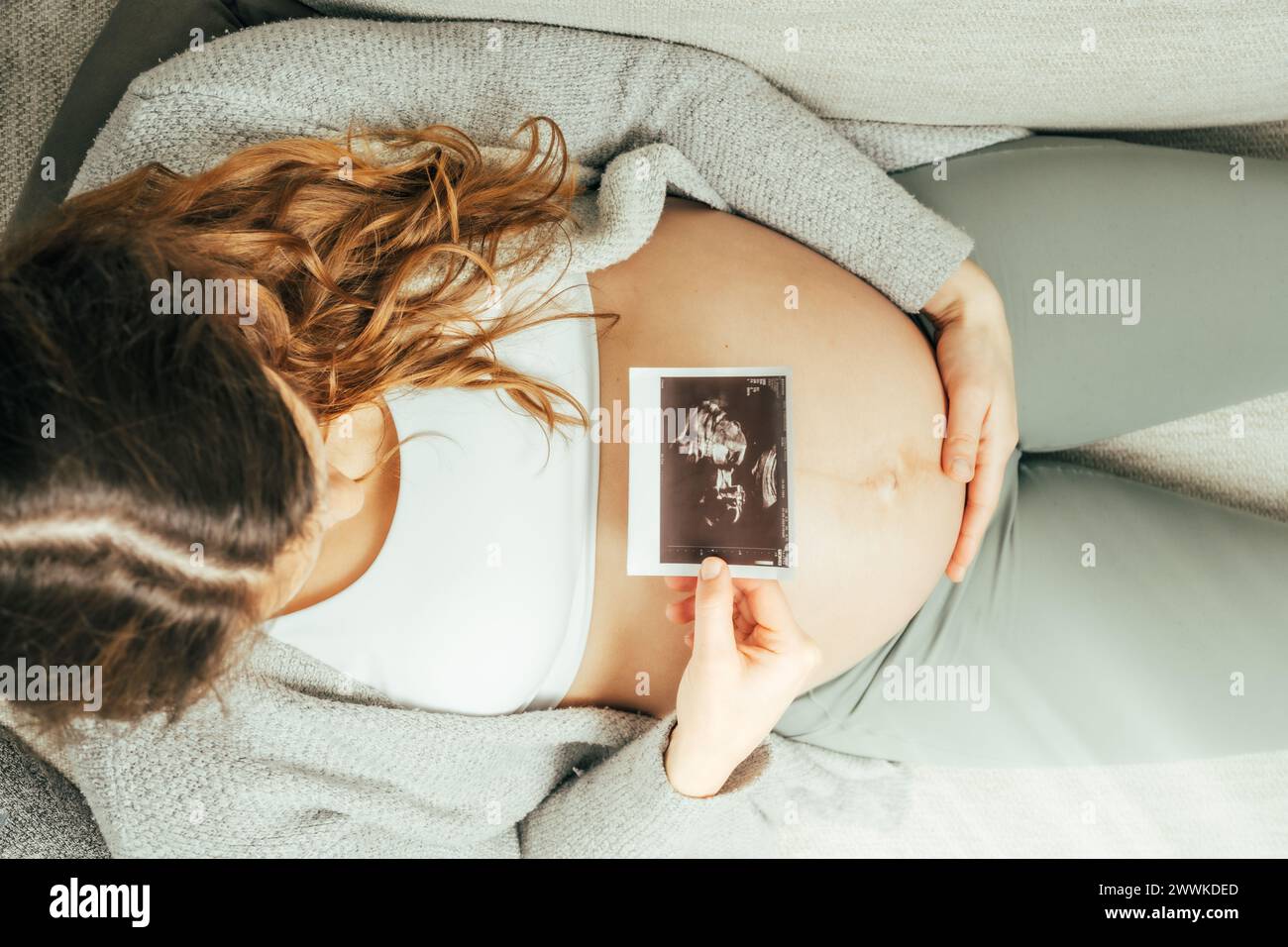 Descrizione: Vista dall'alto di una donna seduta su un divano che guarda un'immagine ecografica del suo bambino nell'ultima fase della gravidanza. Gravidanza, terzo trimestre Foto Stock