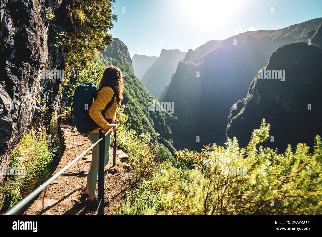 Descrizione: Donna Backpacker godendo di vista panoramica da sotto grande parete di roccia lungo il canale d'acqua a ripida scogliera attraverso la foresta pluviale di Madeira. Levada Foto Stock