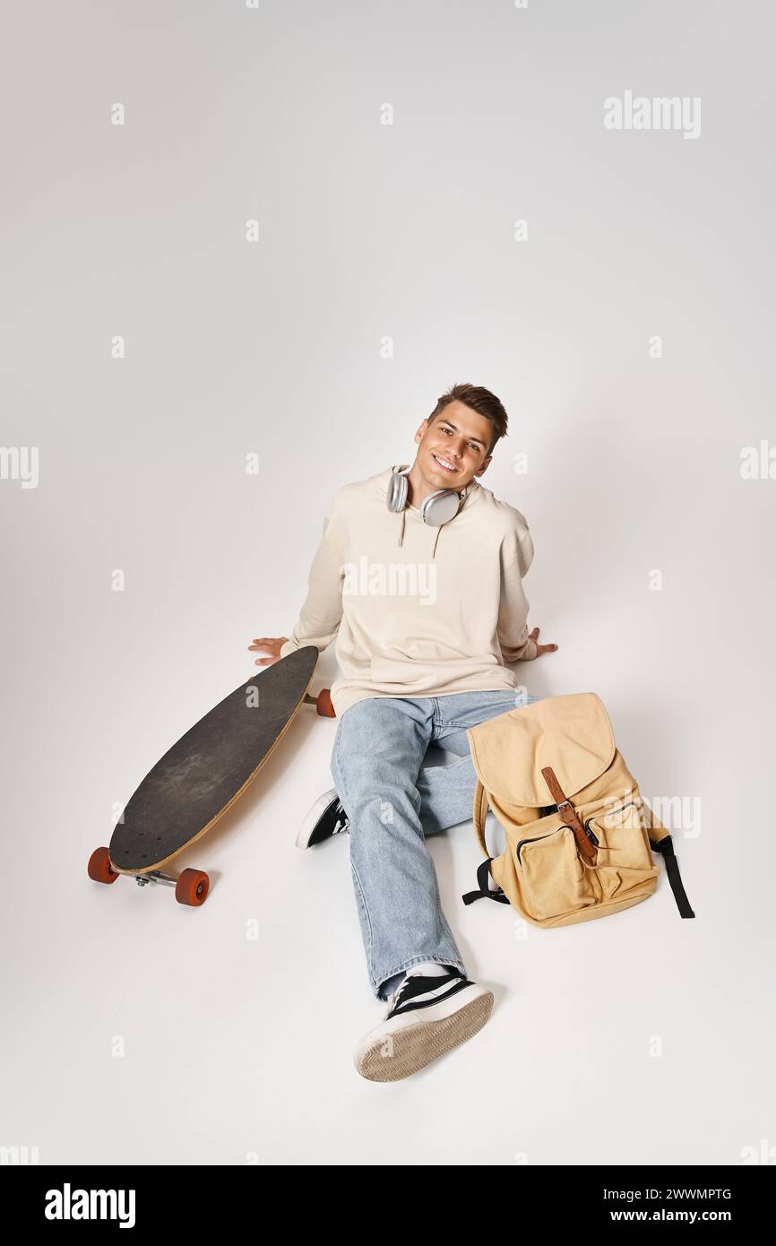 attraente studente con cuffie e abbigliamento casual seduto con zaino e skateboard Foto Stock