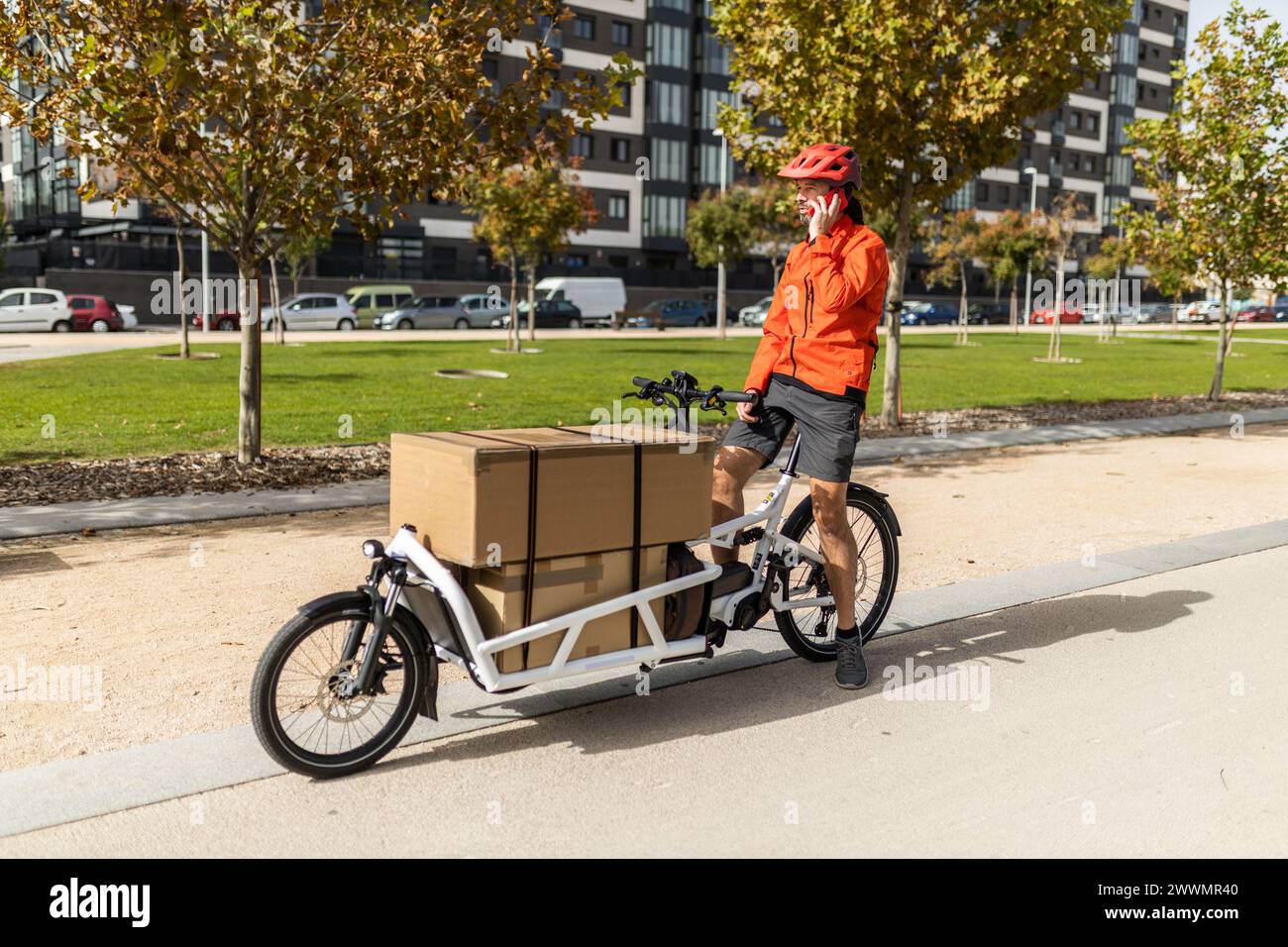 giovane corriere con abbigliamento rosso e casco in bici cargo , percorrendo la pista ciclabile della città per consegnare un pacco, parlando al telefono Foto Stock