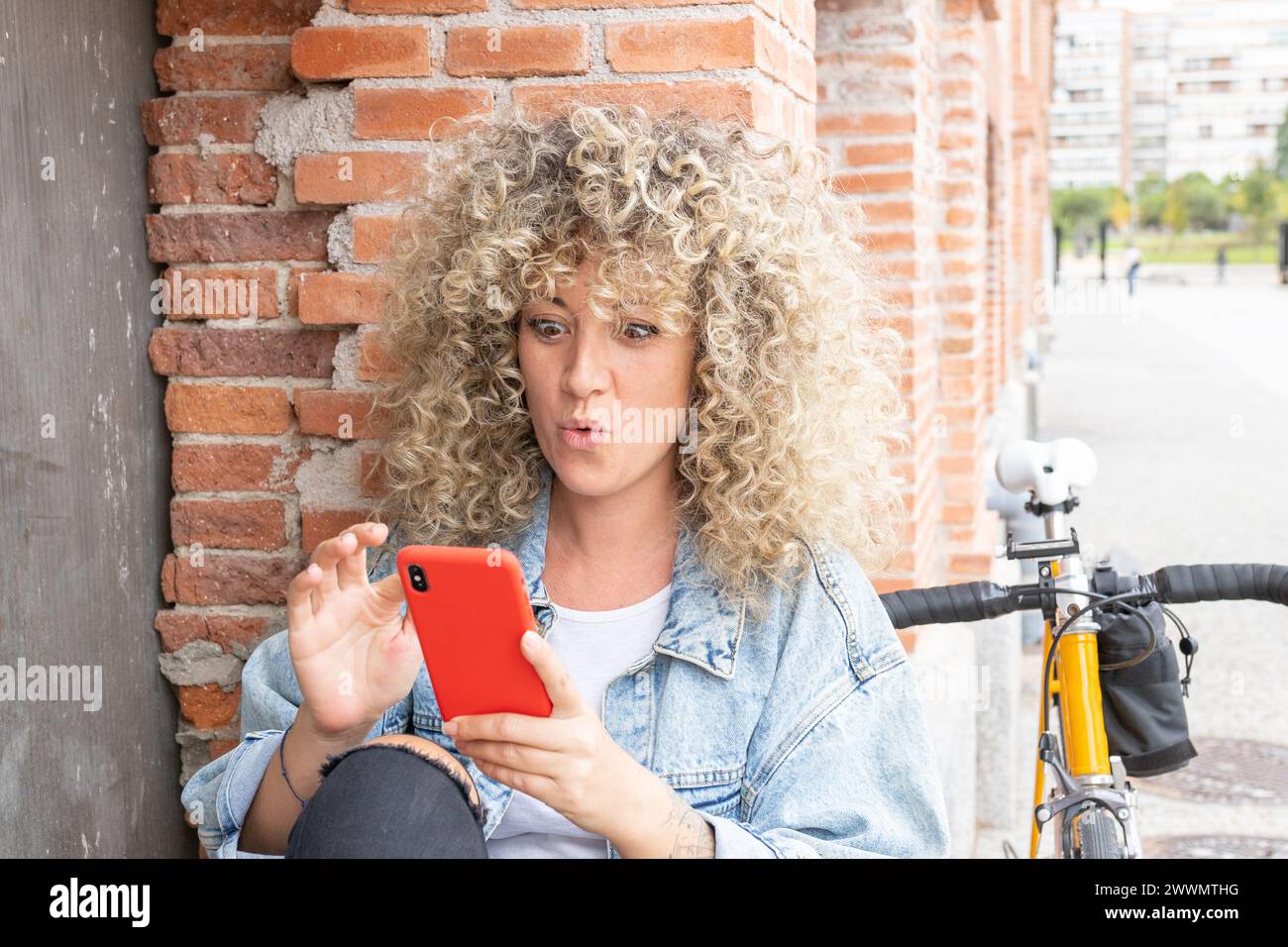 giovane donna caucasica con capelli biondi ricci, sorridente e sorpreso, chiacchierando con il suo smartphone rosso e la sua classica bicicletta gialla Foto Stock