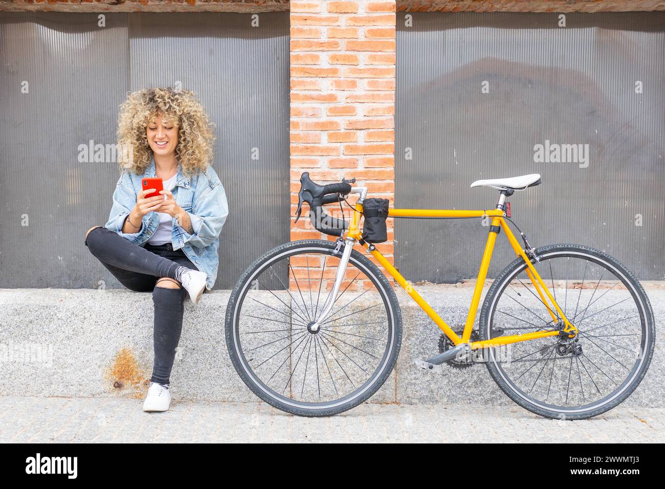 giovane donna caucasica con capelli biondi ricci, sorridente, che chiacchiera con il suo smartphone rosso e la sua classica bicicletta gialla Foto Stock