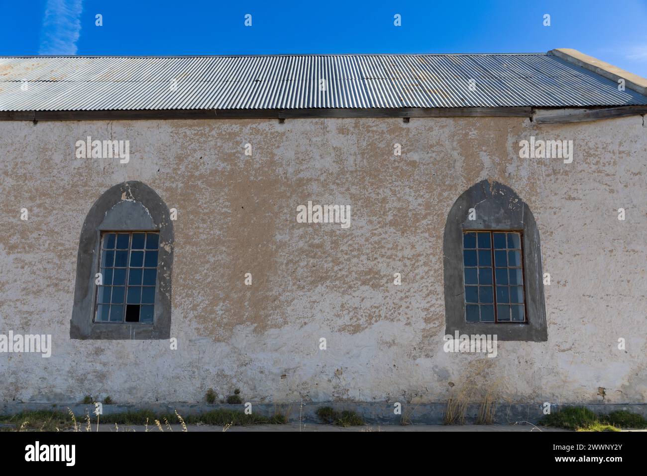 Lato di un vecchio edificio di chiesa abbandonato con tetto in metallo ondulato e due finestre in stile gotico. La vernice bianca si sta staccando dalle pareti. Foto Stock